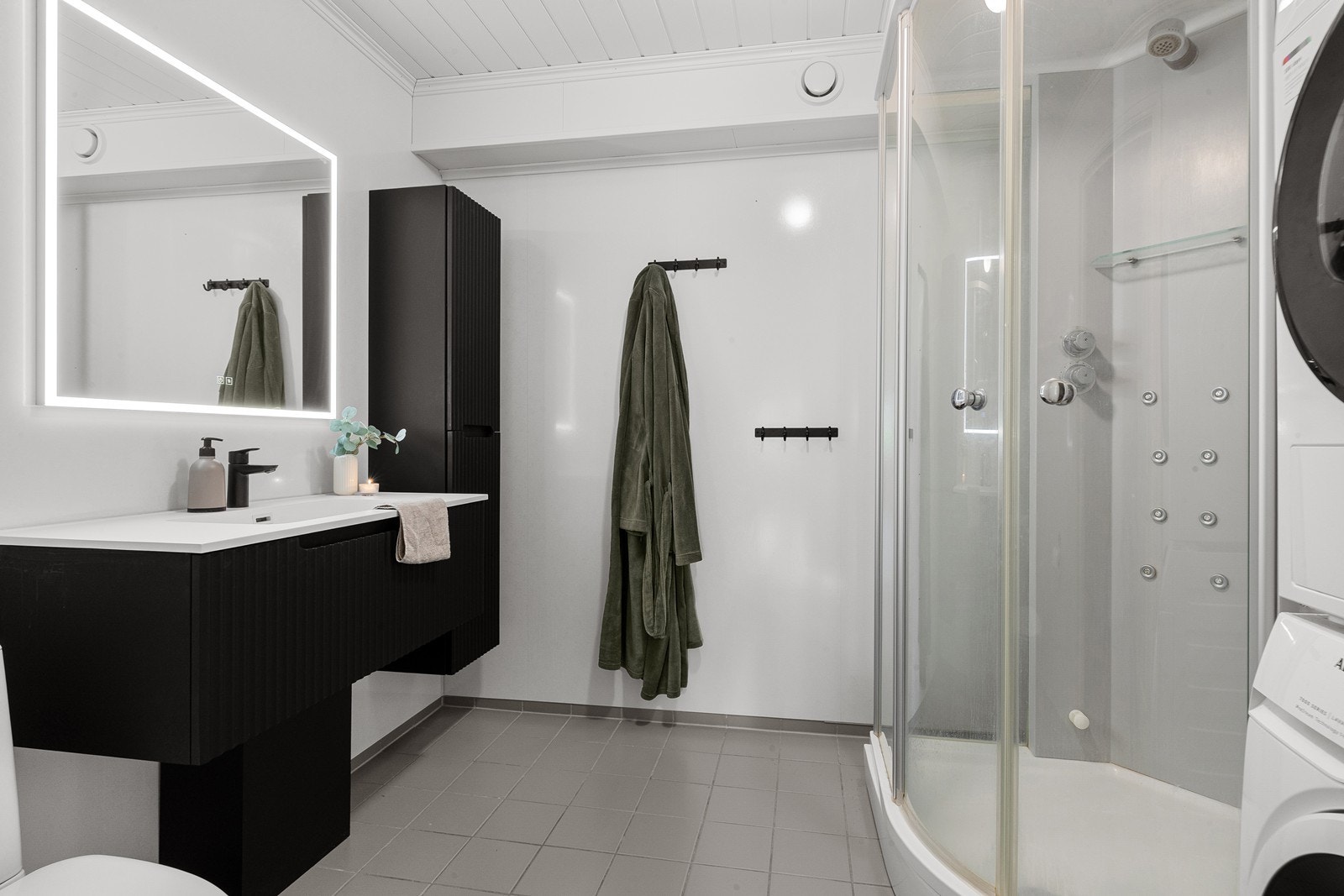Badet har våtromsplater på veggene samt flislagt gulv med elektriske varmekabler. Rommet er innredet med servant, toalett, dusjkabinett og opplegg for vaskemaskin. Galleribilde