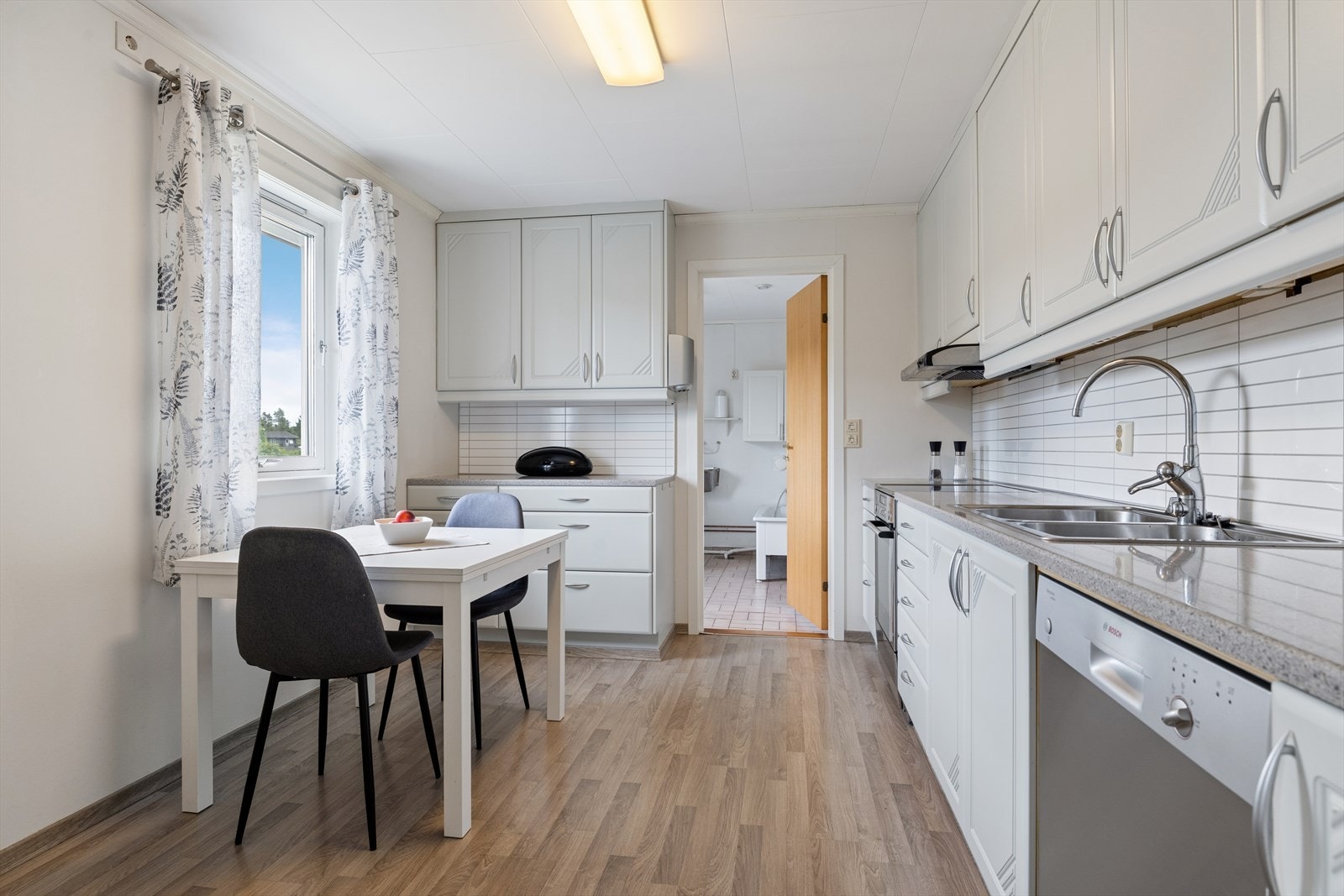 Kjøkkenet har hvite, glatte fronter, benkeplate i steinoptikk og dobbel oppvaskkum i stål. Moderne hvite fliser over benkearealet bidrar til et stilrent uttrykk. Galleribilde