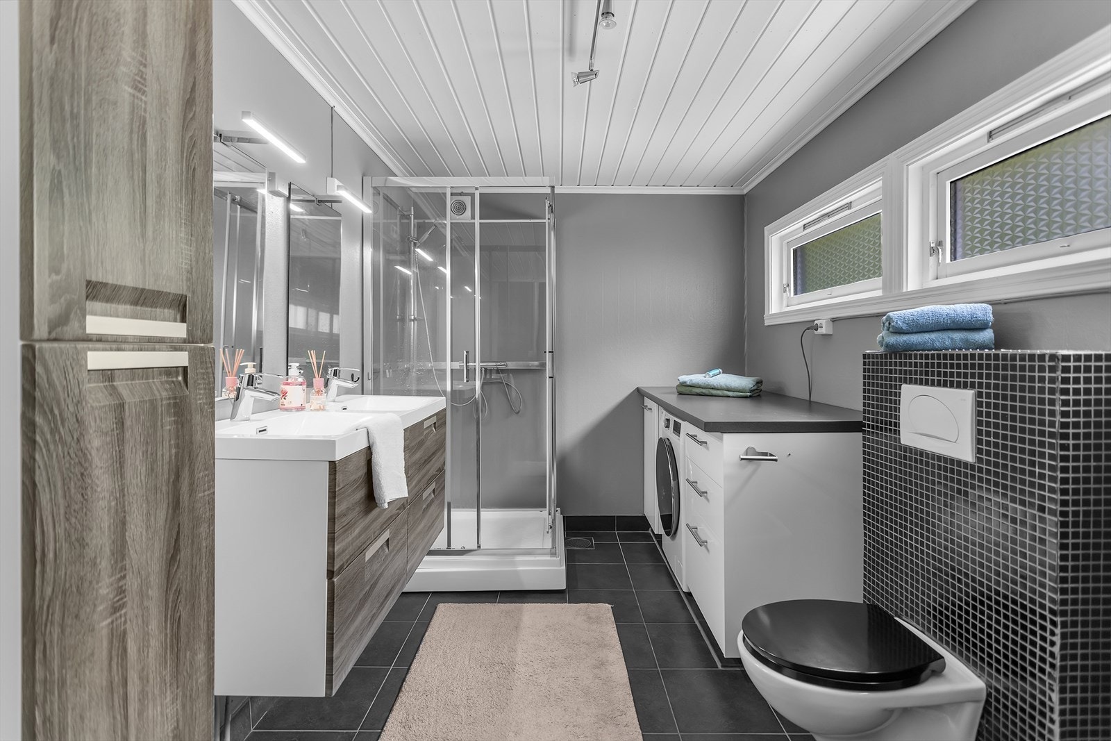 Badet er pent innredet og byr på både stil og praktiske løsninger. Det er utstyrt med et dusjkabinett, dobbelt servant med underskuffer og tilhørende sideskap for ekstra oppbevaringsplass. Galleribilde
