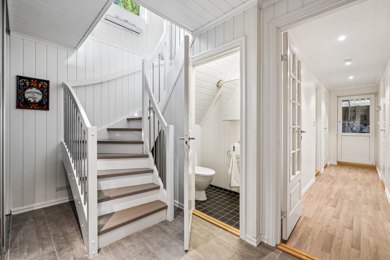 Hall/ gang i 1. etg. med trapp, praktisk toalettrom, garderobe skimtes i venstre bildekant. Til høyre ser vi gangen inn til bad/vaskerom, soverom og bi-utgang. Galleribilde