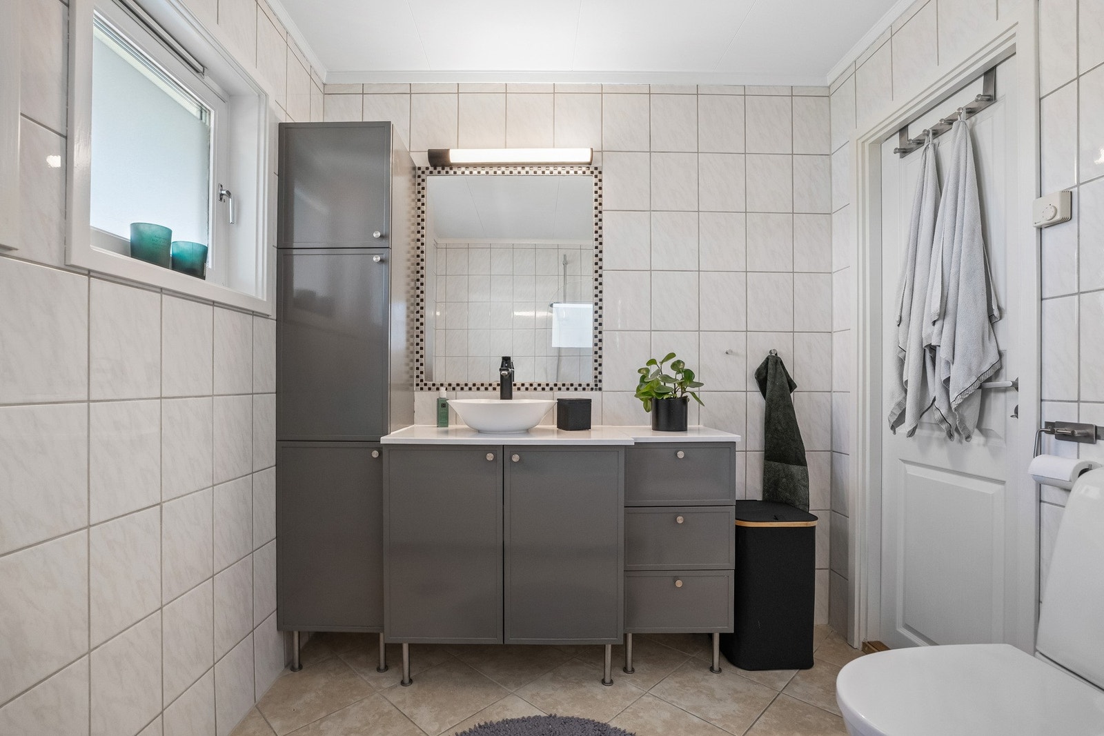 Badet har flislagte overflater og varmekabler i gulvet, samt innredning med skuffer og skap, servant og speil med belysning. Galleribilde