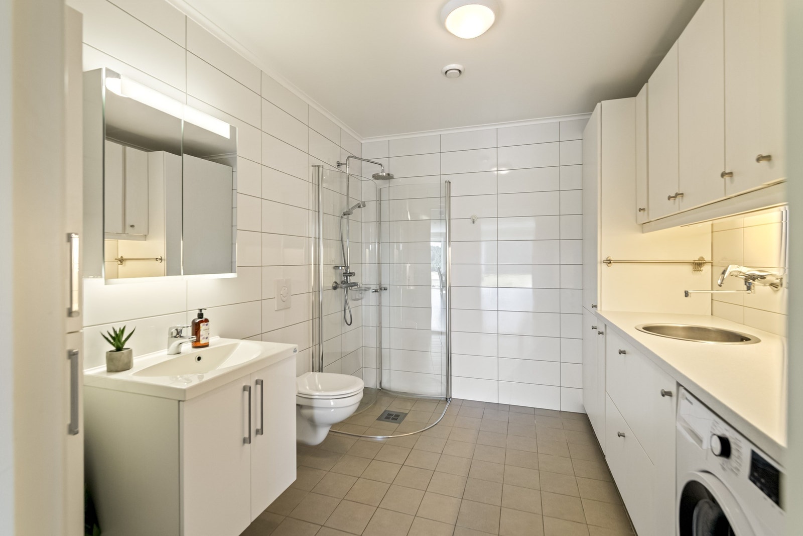 Badet har en funksjonell innredning med rikelig skapplass og praktisk arbeidsbenk. Flislagte gulv og vegger, samt gulvvarme, gir et komfortabelt og helhetlig uttrykk. Galleribilde