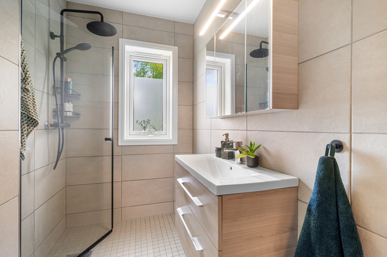 Badet har flislagte gulv og vegger. Oppbevaringsplass i både speilskap og skuffer under servanten. Praktiske innfellbare dusjdører, og både hånd- og takdusj. Galleribilde