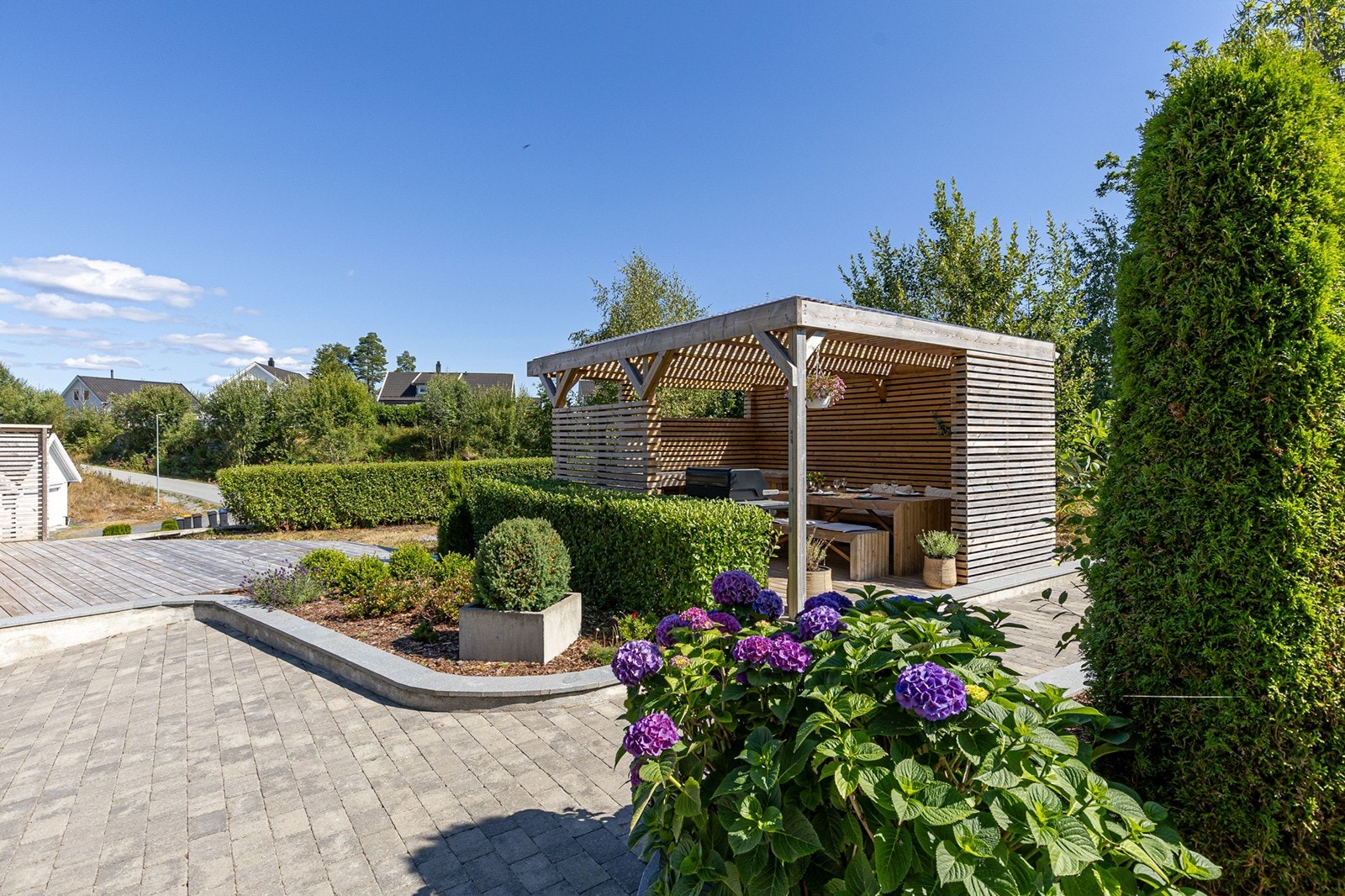 Lun og skjermet pergola med plass til spisegruppe, omkranset av vakre beplantninger og pent opparbeidede uteområder. Galleribilde