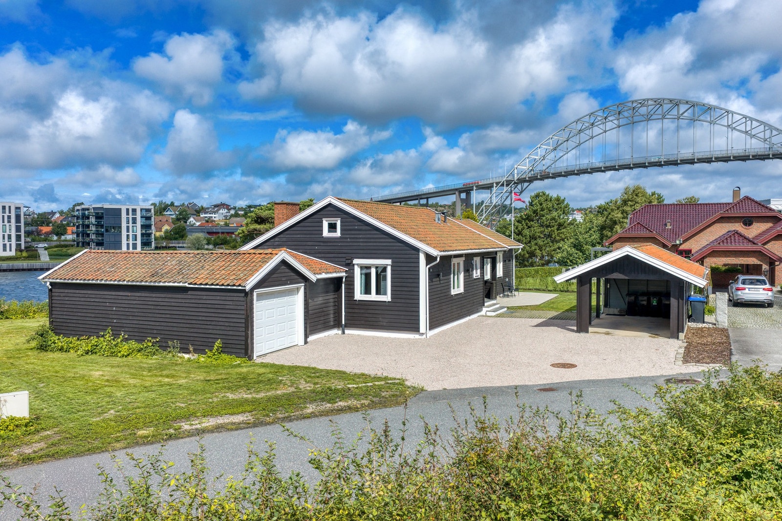 Fra gaten, med garasje, stor gårdsplass og carport. I bakgrunnen den ikoniske Fredrikstadbroa. Galleribilde