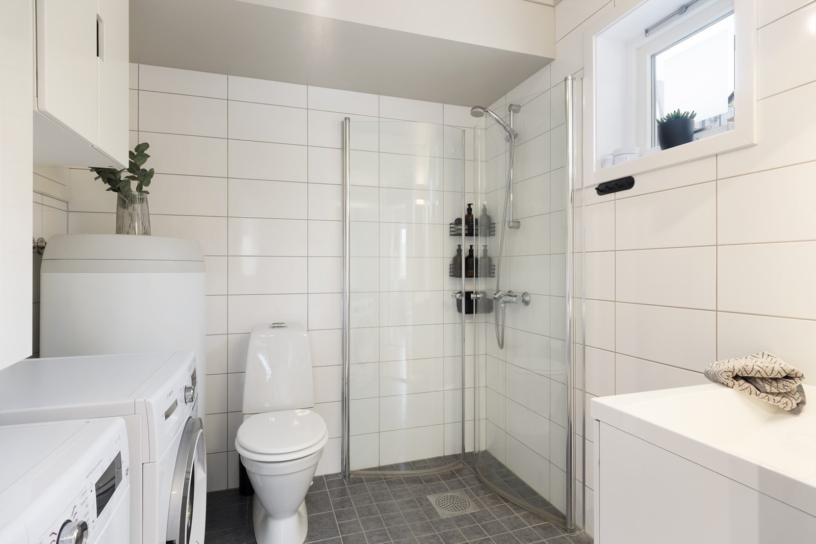 Kombinert bad og vaskerom med flis på golv og veggar. Innreiing består av dusjhjørne med innfellbare glasdører, golvmontert klosett, vask i innredning og opplegg til vaskemaskin. Galleribilde