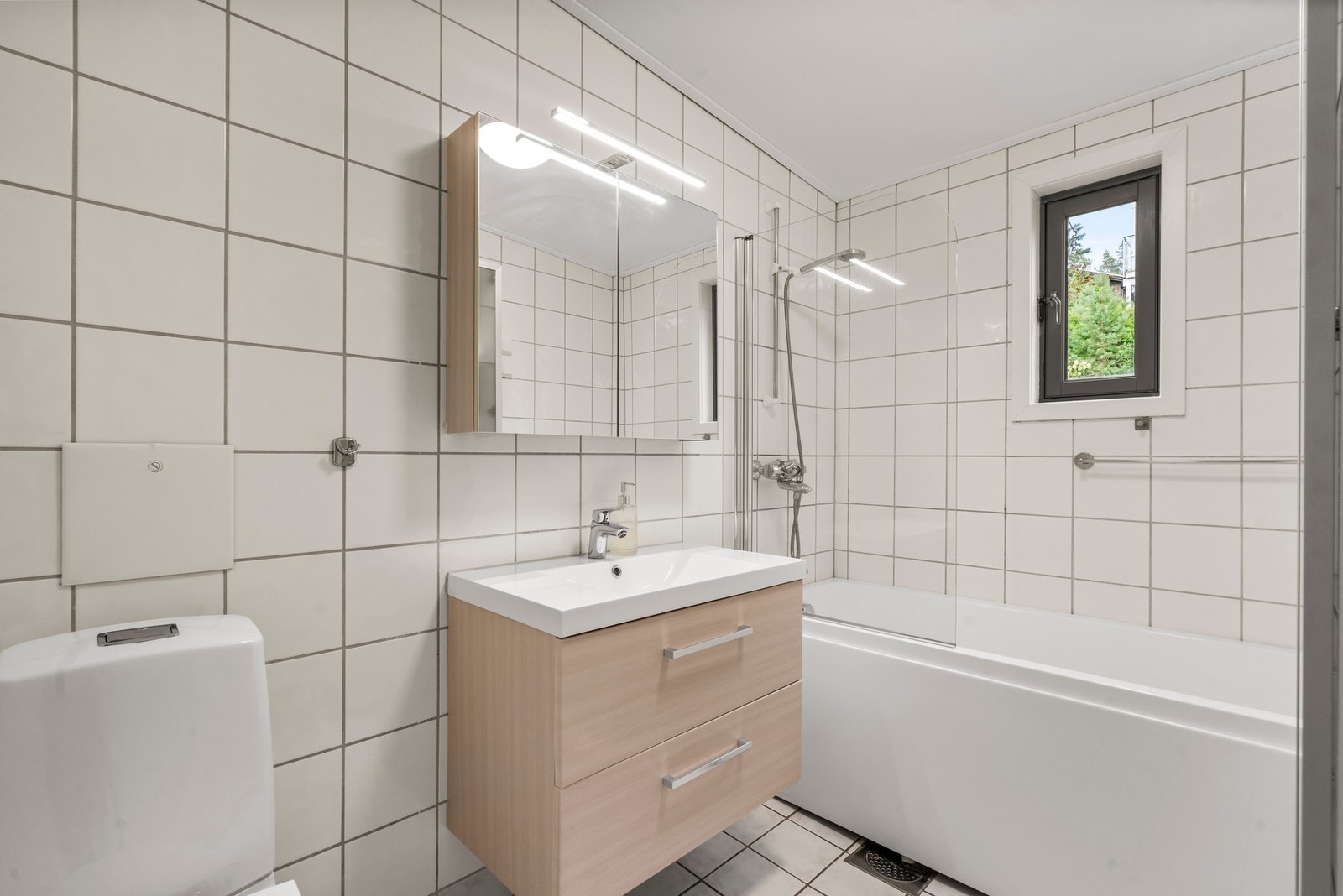 Flislagt bad med gulvvarme, badekar, dusjvegg og speilskap over servanten. Galleribilde