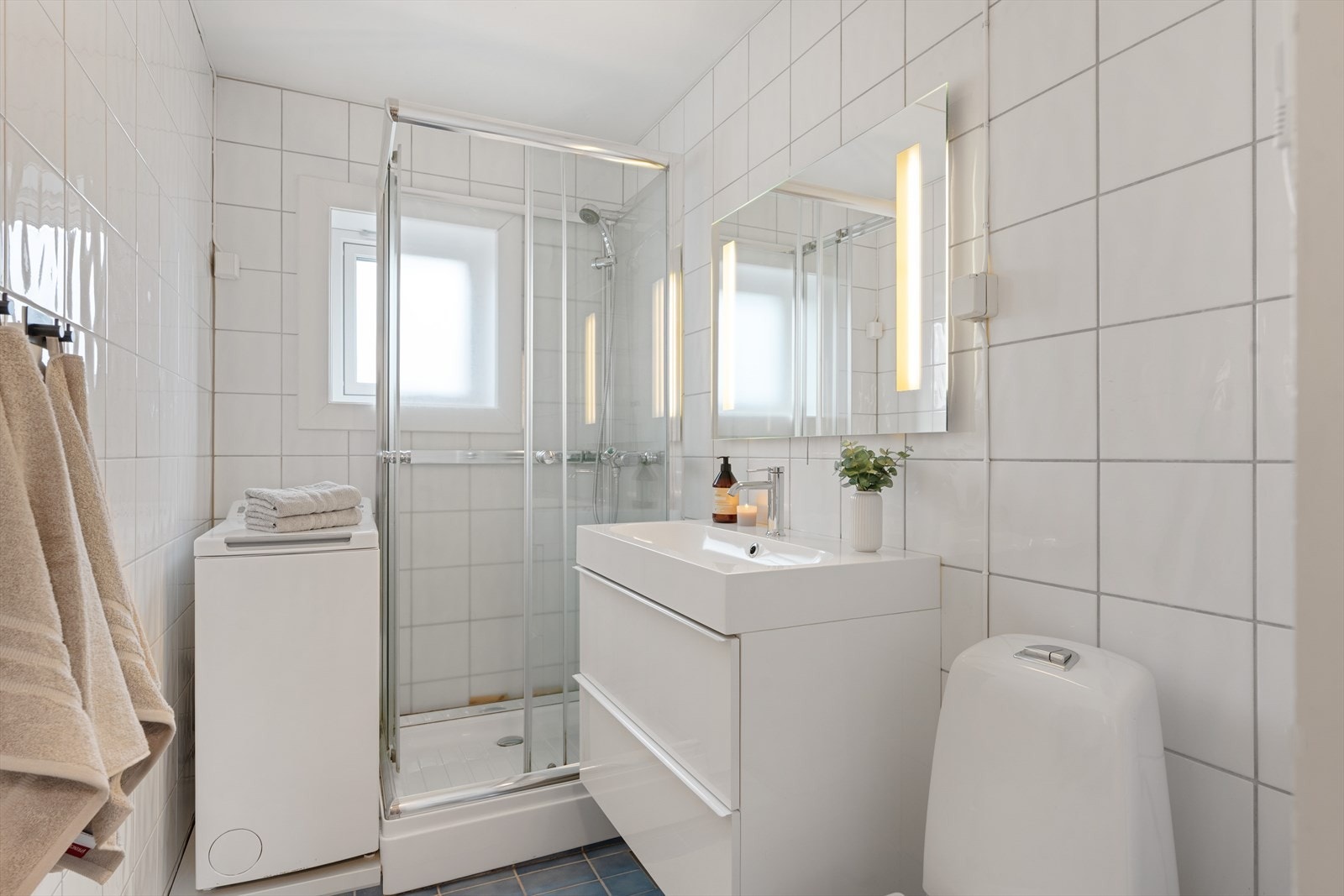 Lyst og flislagt bad med ferdig opplegg for vaskemaskin, praktisk baderomsinnredning og dusjkabinett. Galleribilde
