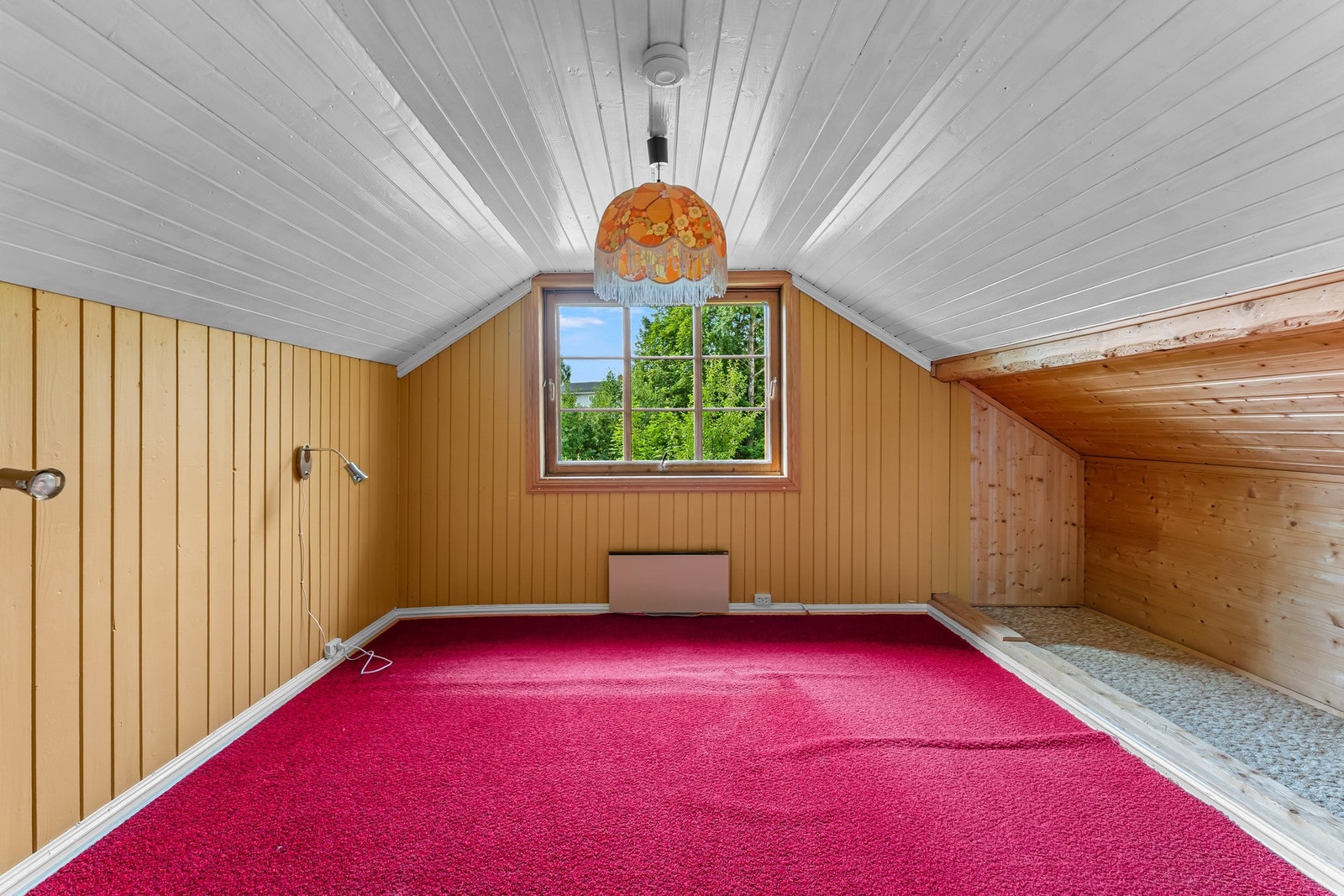 Det neste rommet har vindu i gavlveggen som gir godt dagslys og utsyn mot den grønne vegetasjonen utenfor. Skråtaket gir rommet en lun karakter, mens overflatene gjenspeiler boligens eldre standard. Galleribilde