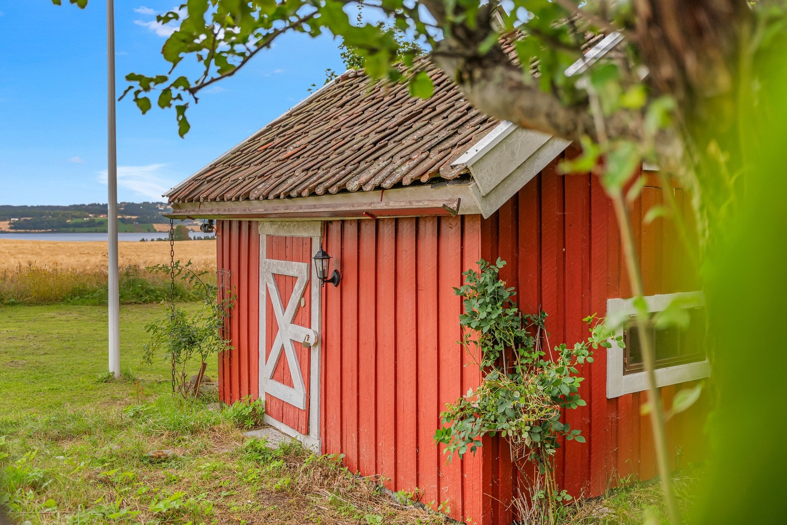 Et sjarmerende punktum for presentasjonen: det røde uthuset med hvite detaljer, klatreplanter og eldre taktekking glir naturlig inn i tunmiljøet, med åkerlandskap og vann som bakteppe. Galleribilde