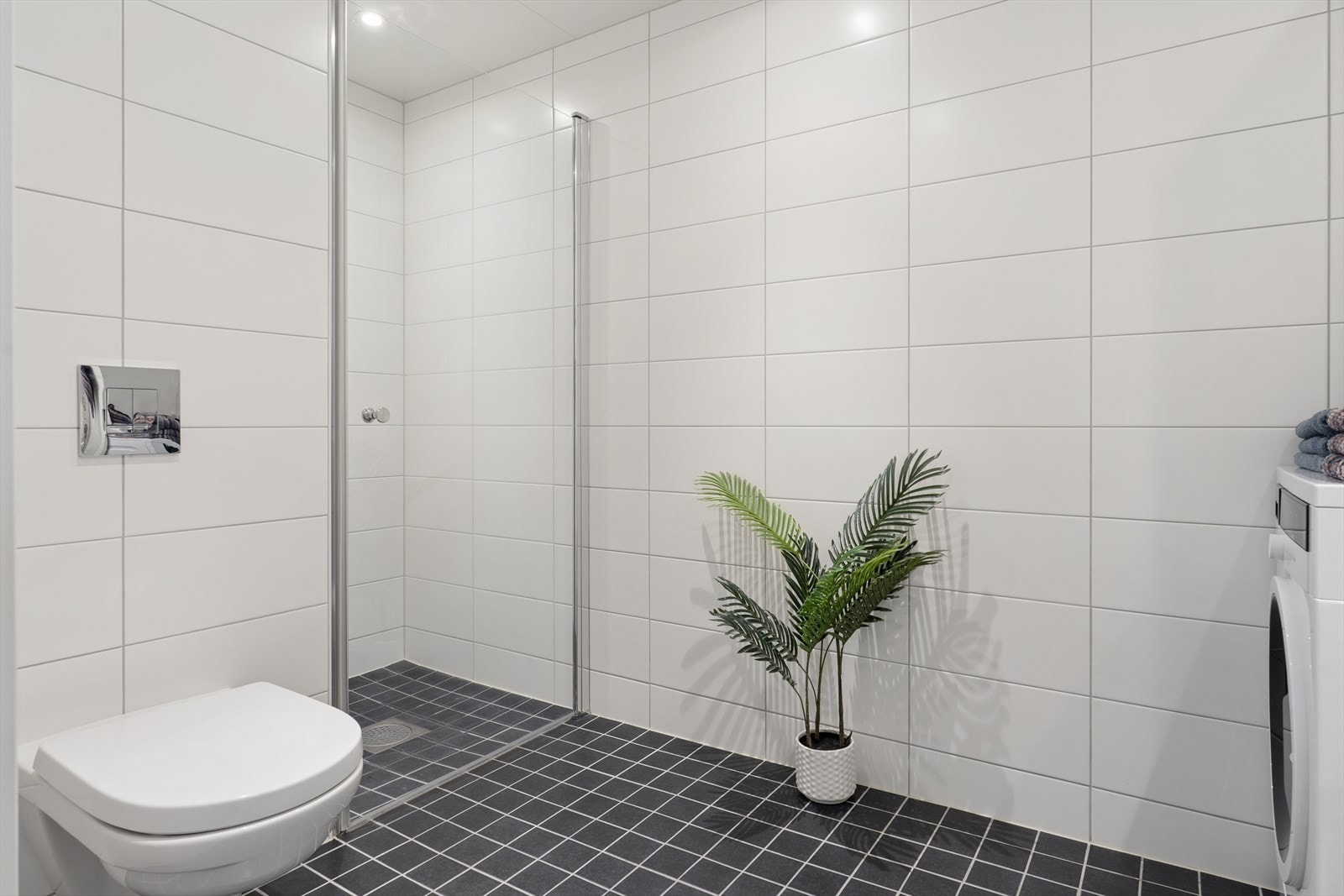 Rommet er innredet med stilren innredning med gode oppbevaringsmuligheter, vegghengt toalett og en praktisk dusjnisje med glassdør. Det er opplegg for vaskemaskin, pent integrert i et skap rundt maskinen, noe som gir et ryddig og gjennomført uttrykk. Galleribilde