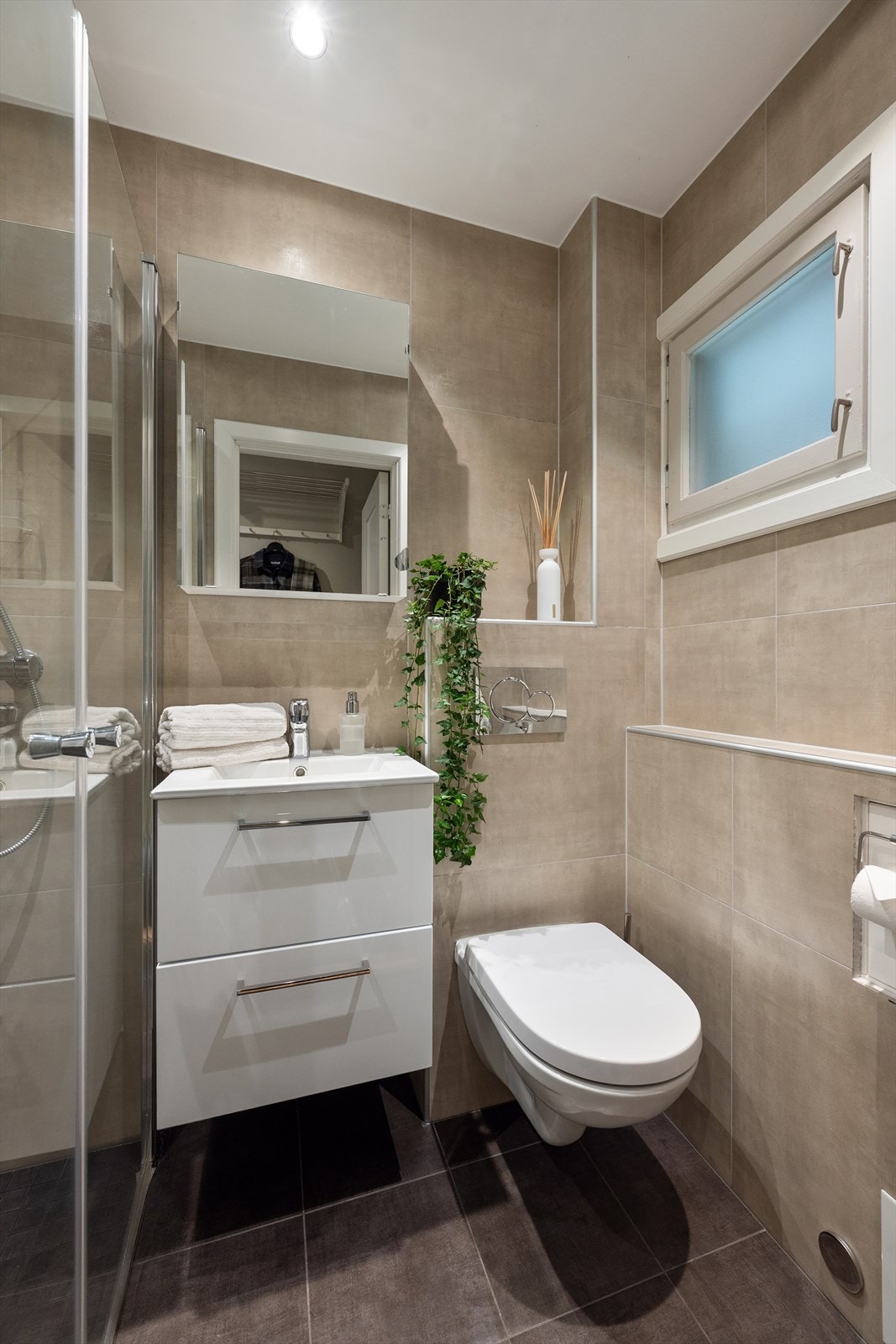 Badet er flislagt med pene beige fliser samt innredet med dusj med dørvegg, servant i skuffeinnredning, speilskap med innredning og vegghengt toalett. Galleribilde