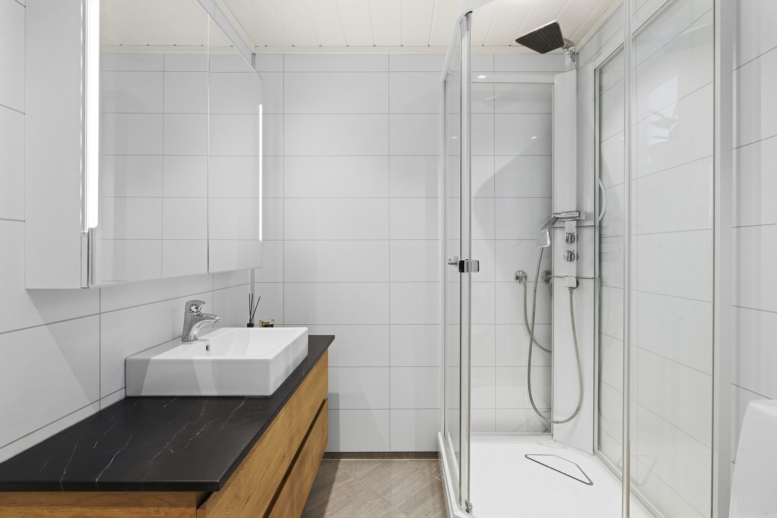 Badet inneholder toalett, en bred baderomsinnredning med overliggende servant, skapspeil med belysning, samt et dusjkabinett. Galleribilde