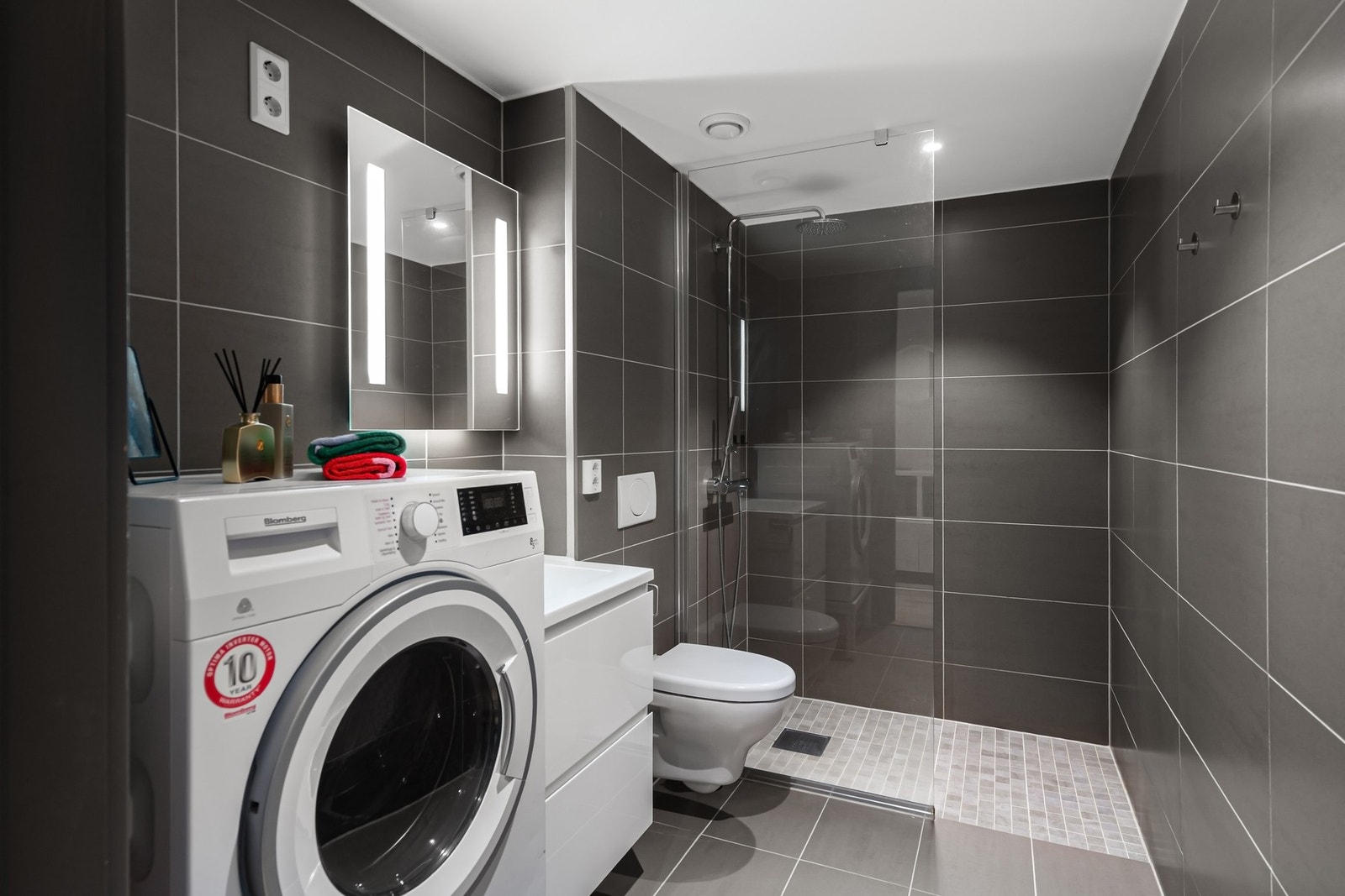 Badet har et moderne uttrykk med flislagte vegger og gulv. Rommet er innredet med åpen dusjløsning, vegghengt toalett og servant med oppbevaring. Galleribilde