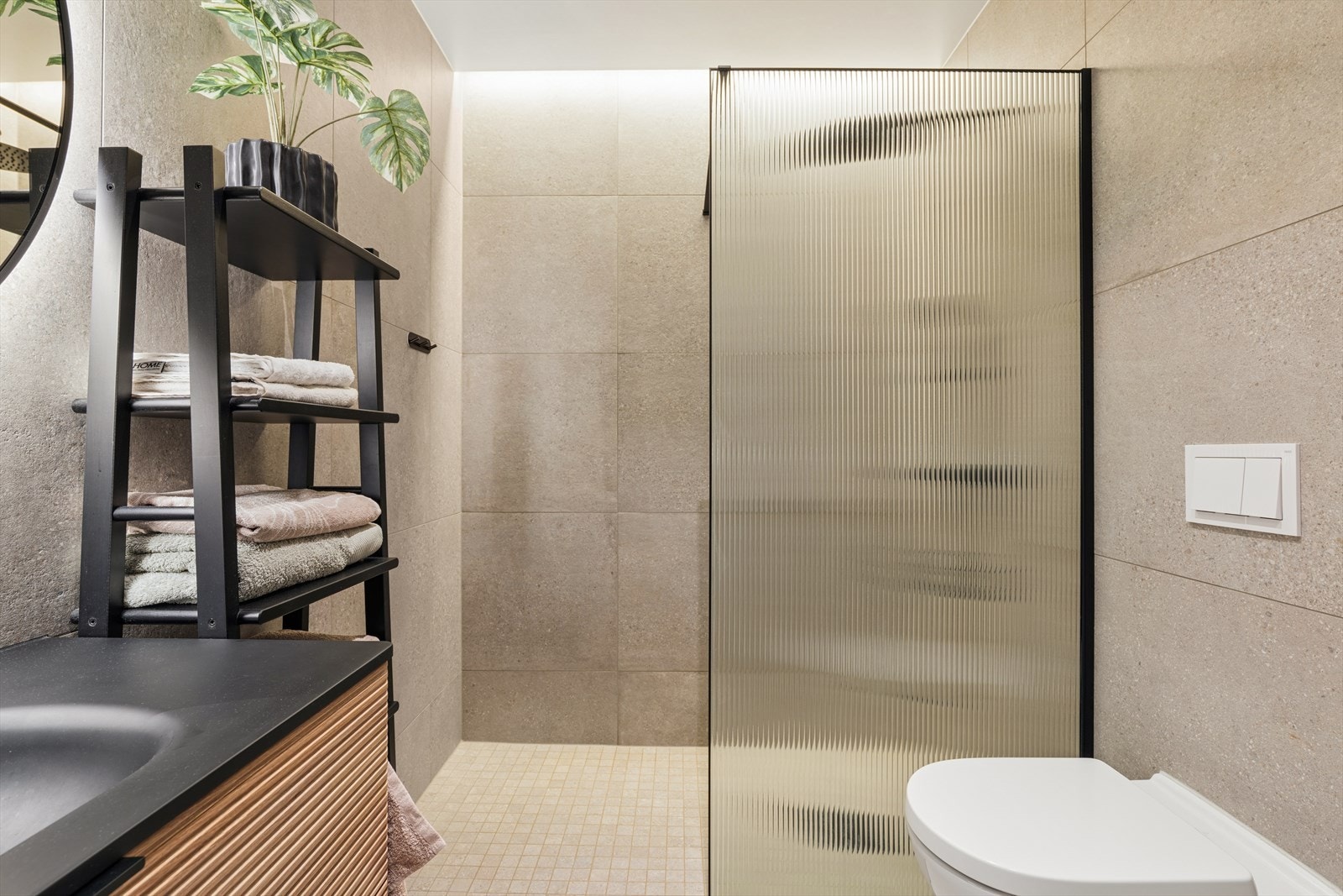 Lekkert bad med fliser på vegger og gulv med gulvvarme. Badet har innredning med servant, dusjhjørne og veggmontert toalett. Galleribilde
