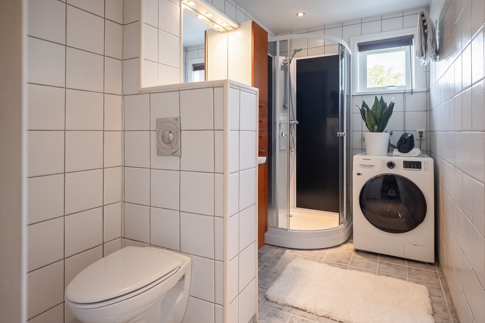Badet er utstyrt med praktisk baderomsinnredning, speil med integrert belysning, vegghengt toalett og dusjkabinett. Galleribilde