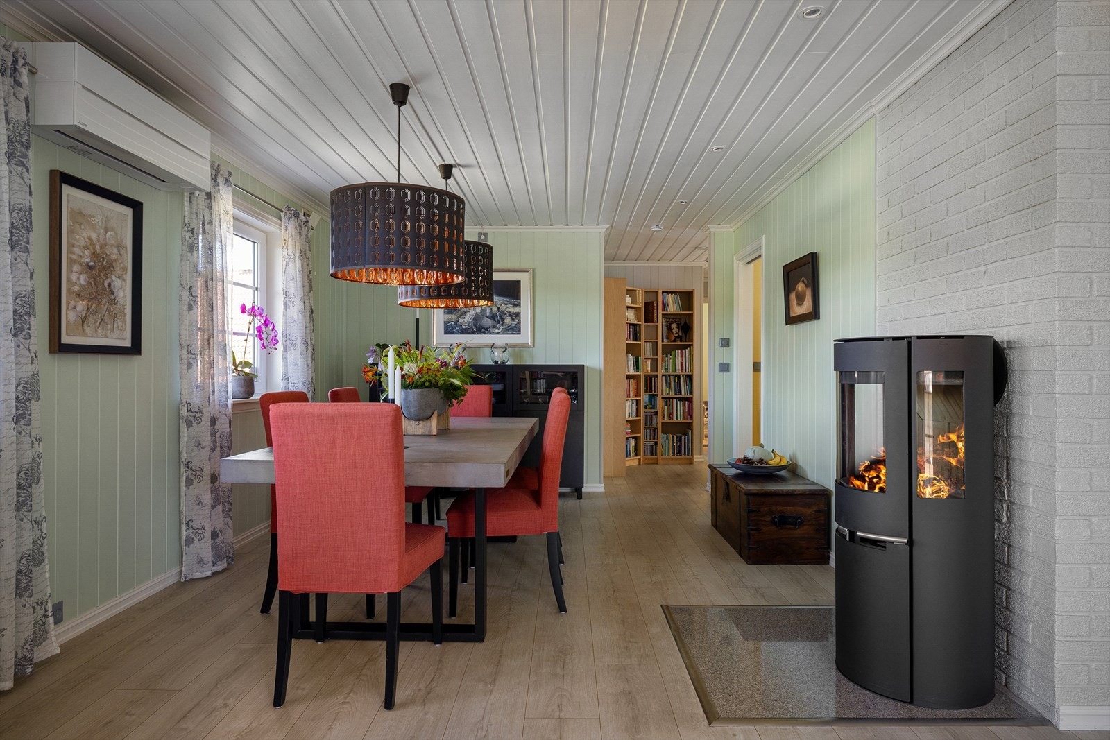 De fleste veggene er kledd med malt panel, i likhet med taket, mens noen er tapetserte. Gulvets slitesterke laminat flyter fint og helhetlig innover det store stuerommet. På veggen henger en varmepumpe (2021) som avlaster og supplerer peisovnen (2023). Galleribilde