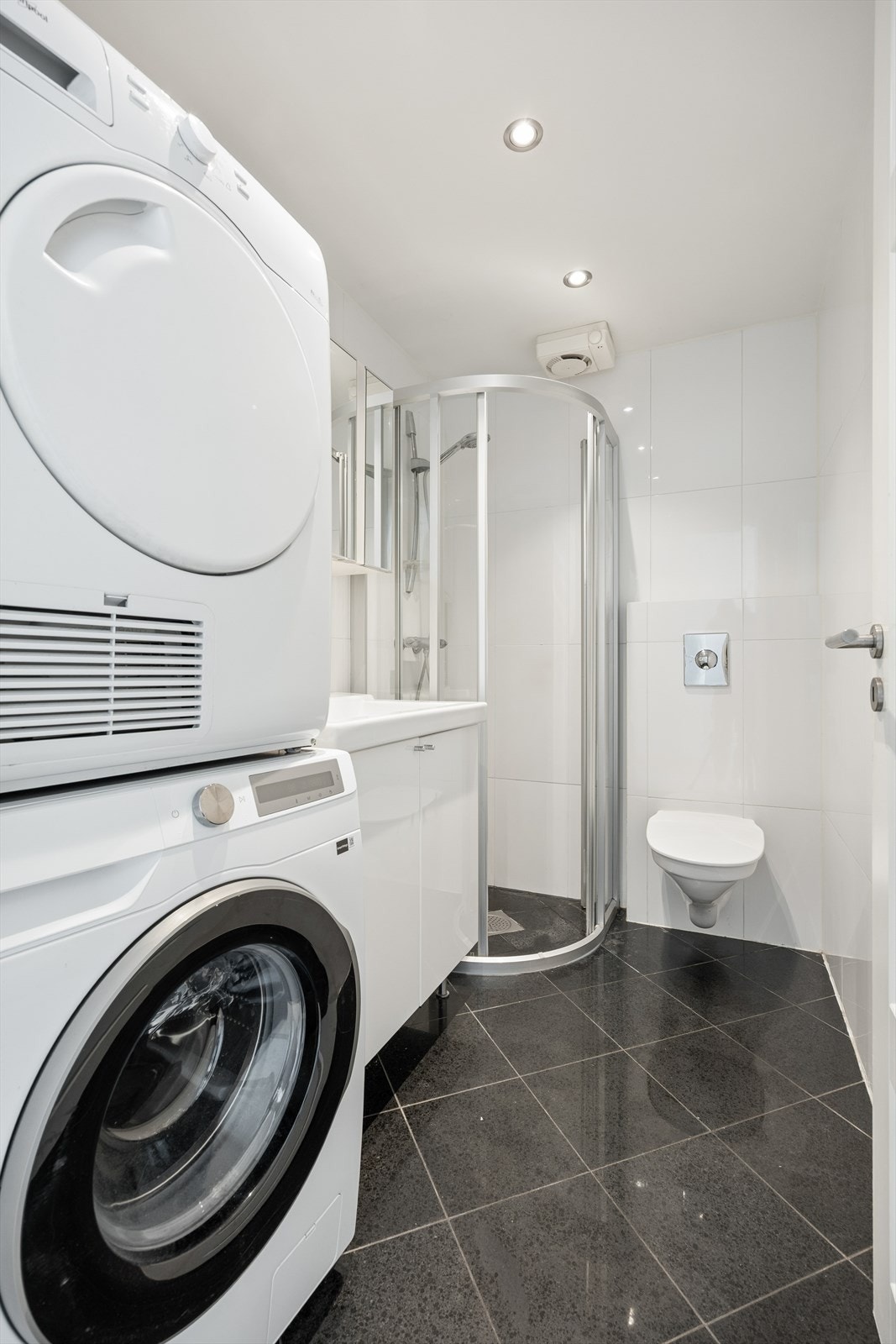 Badet har flislagt gulv, opplegg for vaskemaskin og tørketrommel, dusjsone, vegghengt toalett og lys baderomsinnredning med skuffeløsninger for oppbevaring. Galleribilde