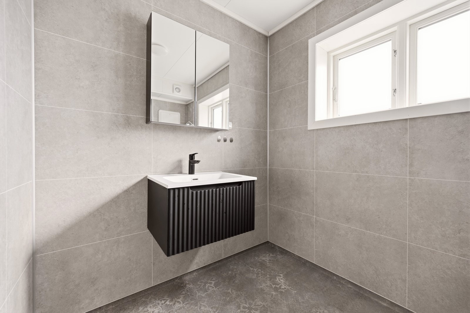 Badet er utstyrt med vegghengt toalett, dusjkabinett og servant nedfelt i skuff med mørk innredning. Speilskap over servanten, og vinduer med frostet glass på langveggen. Galleribilde