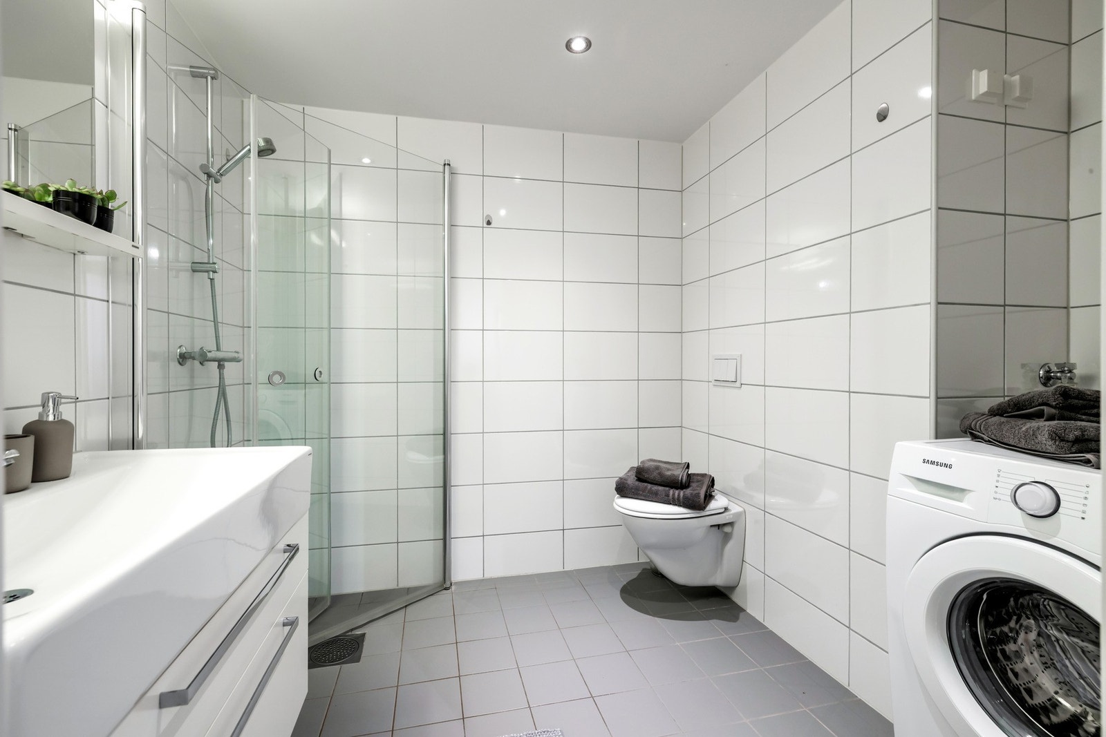 Badet er romslig og har et lyst uttrykk med flislagte vegger og gulv. Rommet er innredet med vegghengt toalett, servantinnredning med god oppbevaringsplass og dusjhjørne med glassdører. Badet har også praktisk opplegg til vaskemaskin. Galleribilde