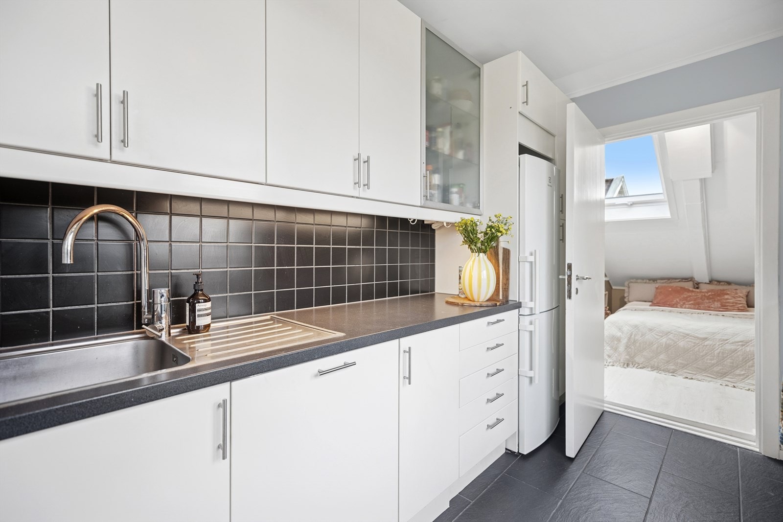 Stilrent kjøkken med glatte fronter, belysning under overskap, laminert benkeplate med nedfelt kum i rustfritt stål, fliser på vegg over benkeskap, samt opplegg for oppvaskmaskin. Galleribilde