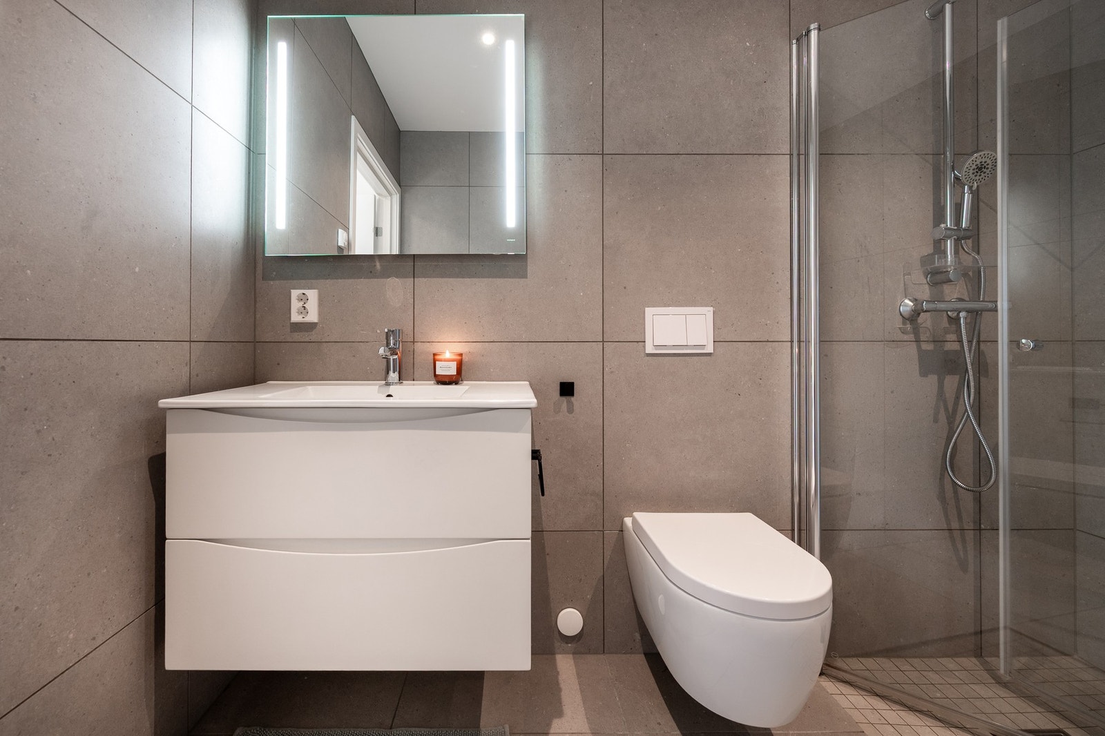 Badet har gulvvarme og er innredet med en hvit, vegghengt servantinnredning med skuffer, speil med integrert belysning, vegghengt toalett og dusjnisje med glassdører. Galleribilde