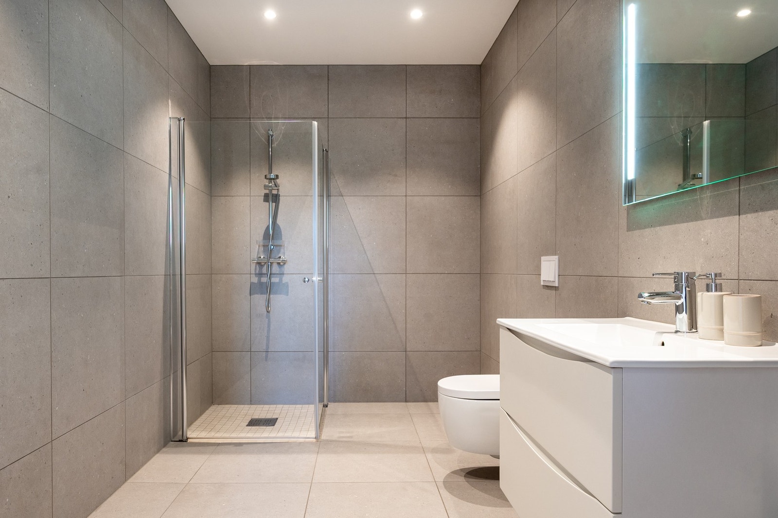 Badet ved gangen har flislagte gulv og vegger i gråtoner, gulvvarme og downlights i himlingen. Galleribilde