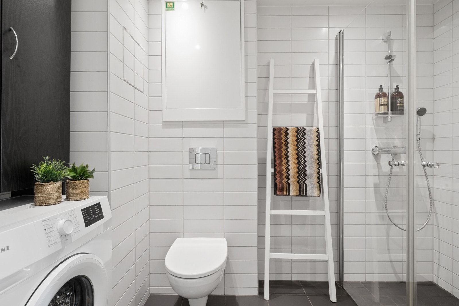 Videre er badet utstyrt med vegghengt toalett, opplegg for vaskemaskin og et dusjhjørne med innfellbare glassdører. Galleribilde