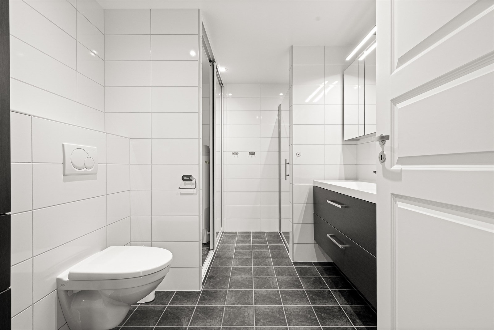 Lyst og moderne flislagt baderom med pent baderomsmøbel, speilskap, vegghengt toalett, dusjnisje og downlights i himling. Galleribilde