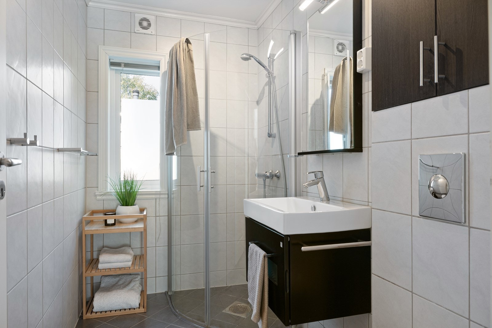 Badet fremstår tidløst med fliser på gulv og vegger og er utstyrt med dusjhjørne, vegghengt toalett, servant i seksjon. Galleribilde