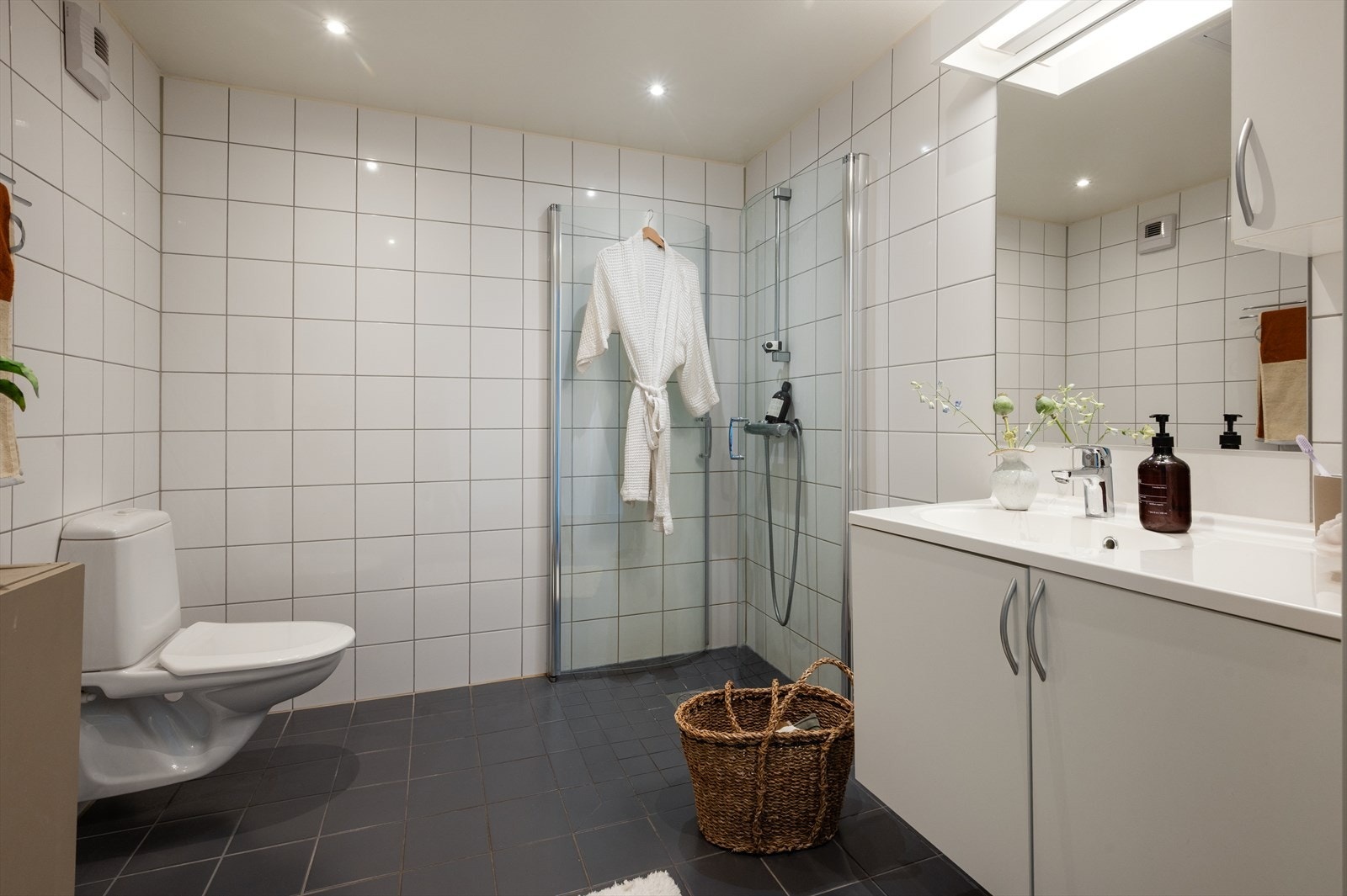 Badet har fliser på vegger og gulv, vegghengt toalett, servantinnredning og dusjhjørne med svingbare glassdører. Galleribilde