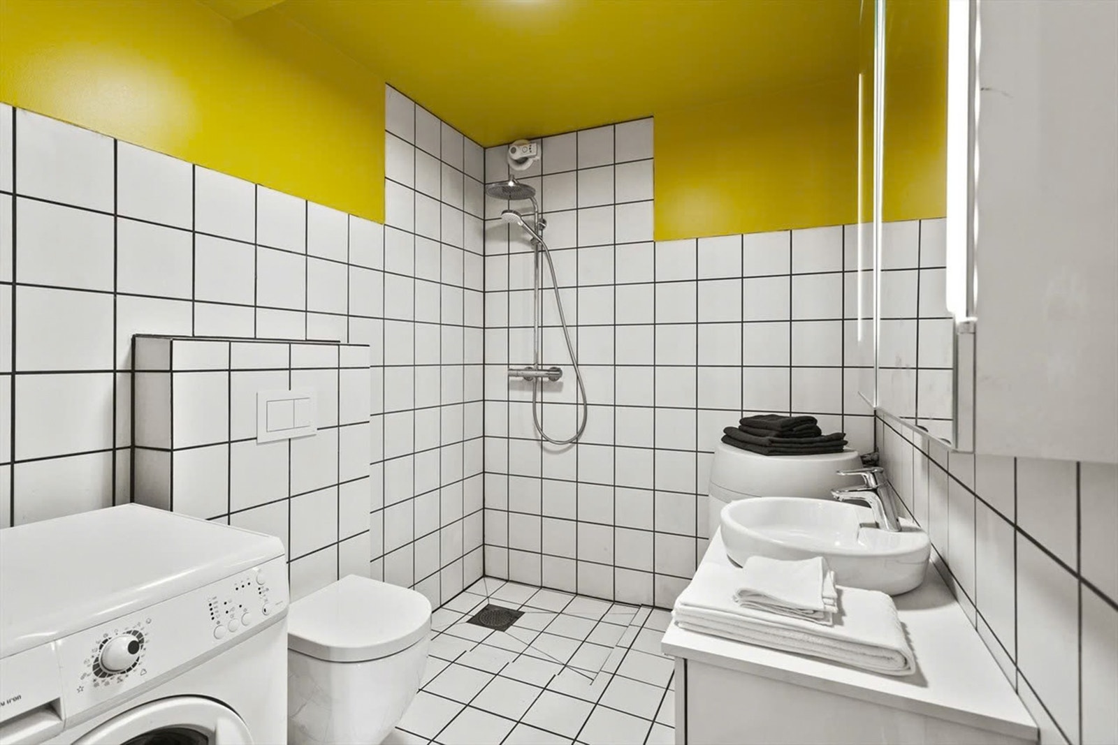 Badet i leiligheten har gulmalte, glatte flater, hvite fliser på veggene og matchende fliser på gulv med gulvvarme. Galleribilde