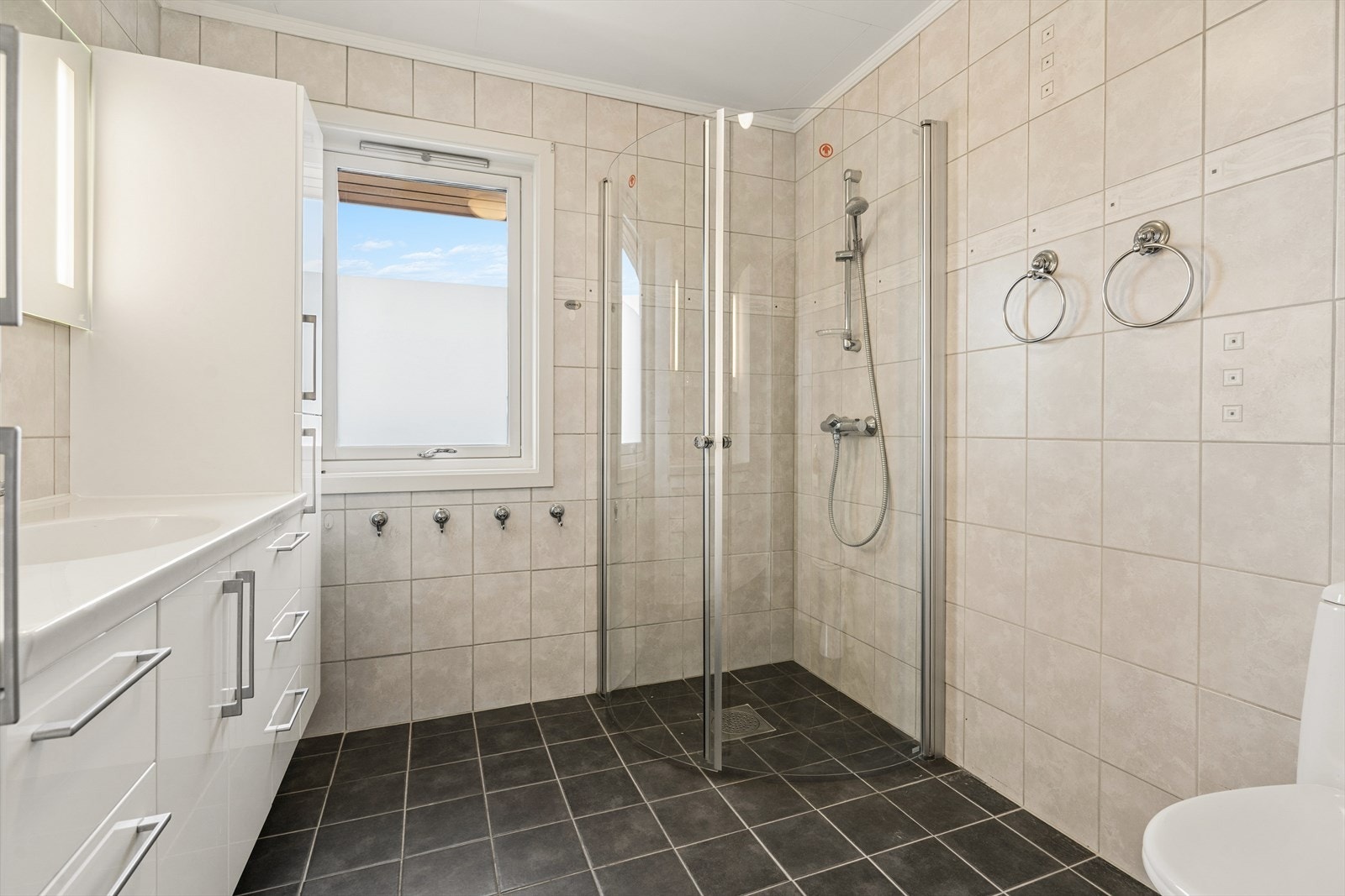 Badet i 1. etasje har flislagte overflater på gulv og vegger. Rommet er innredet med servant med underskap, speil med tilhørende belysning, dusjvegger og gulvmontert toalett. Galleribilde