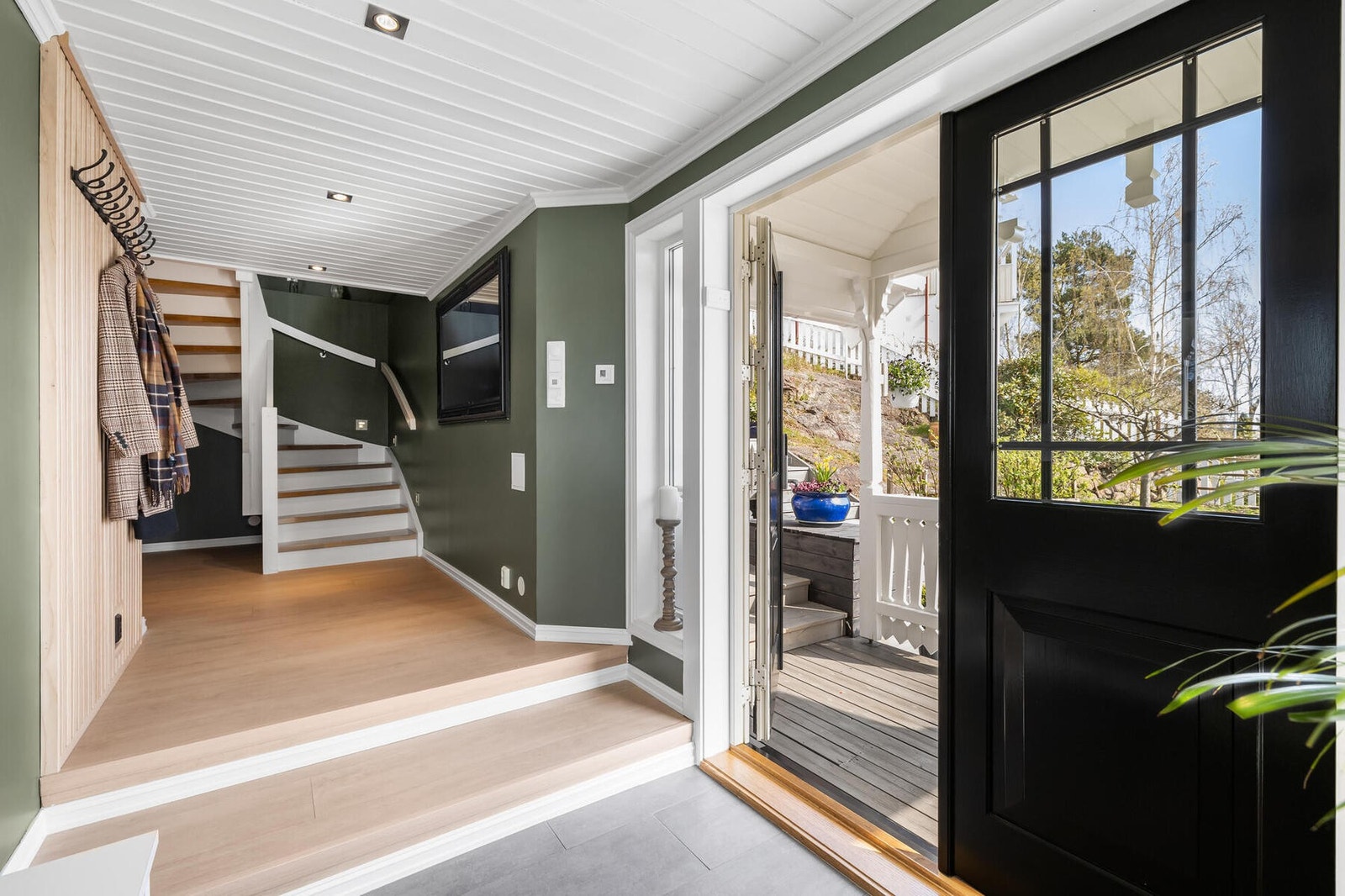Innenfor en tofløyet, sort ytterdør, blir vi ønsket velkommen inn i en stilfull entré med grønnmalte vegger, downlights og grå fliser på gulv med gulvvarme. Galleribilde