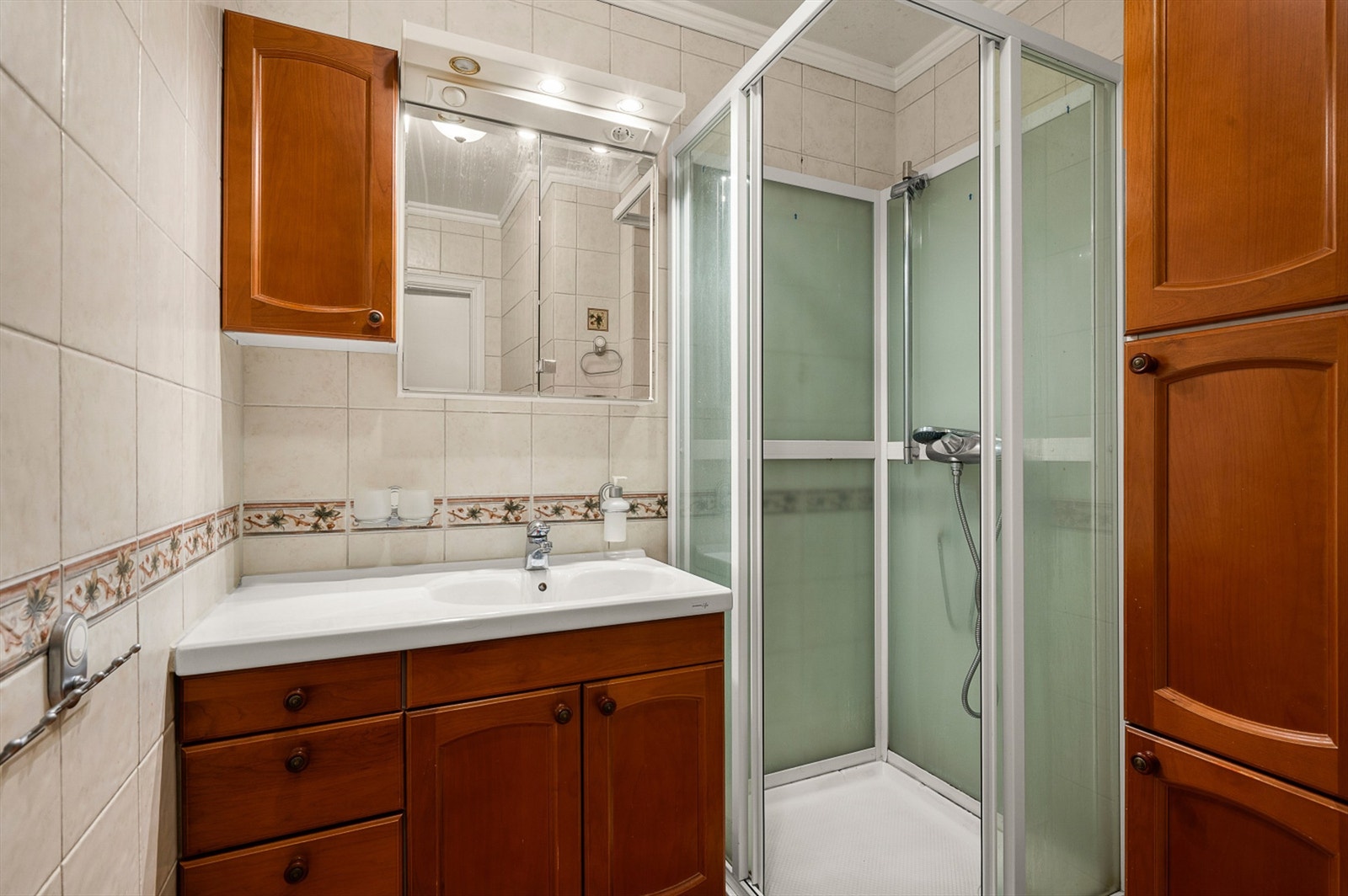 Badet måler 3 kvm og er innredet med flislagte flater, servantskap med overskap og speil med lys, sideskap og dusjkabinett. Galleribilde