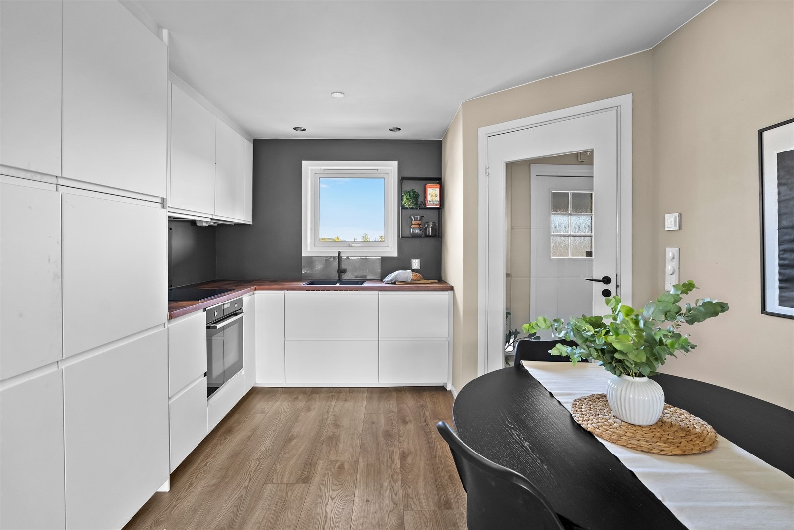 Kjøkkeninnredning med malte glatte fronter, valnøtt benkeplater m/sponkjerne, underlimt oppvaskkum, ventilator, plate over benk og belysning under overskap. Integrerte hvitevarer bestående av oppvaskmaskin, kjøl/fryseskap, stekeovn og induksjonstopp. Galleribilde