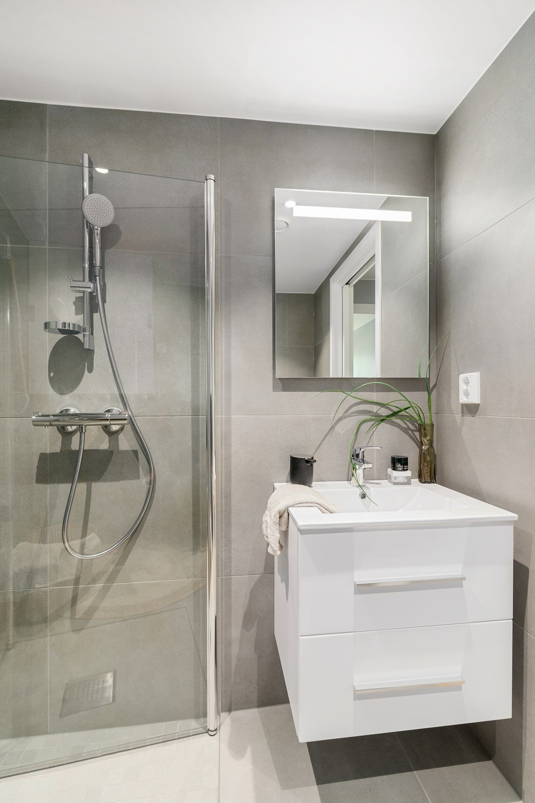 Badet er innredet med servant i hvit skuffeseksjon, speil, vegghengt toalett og romslig dusjsone med glassdører. Galleribilde