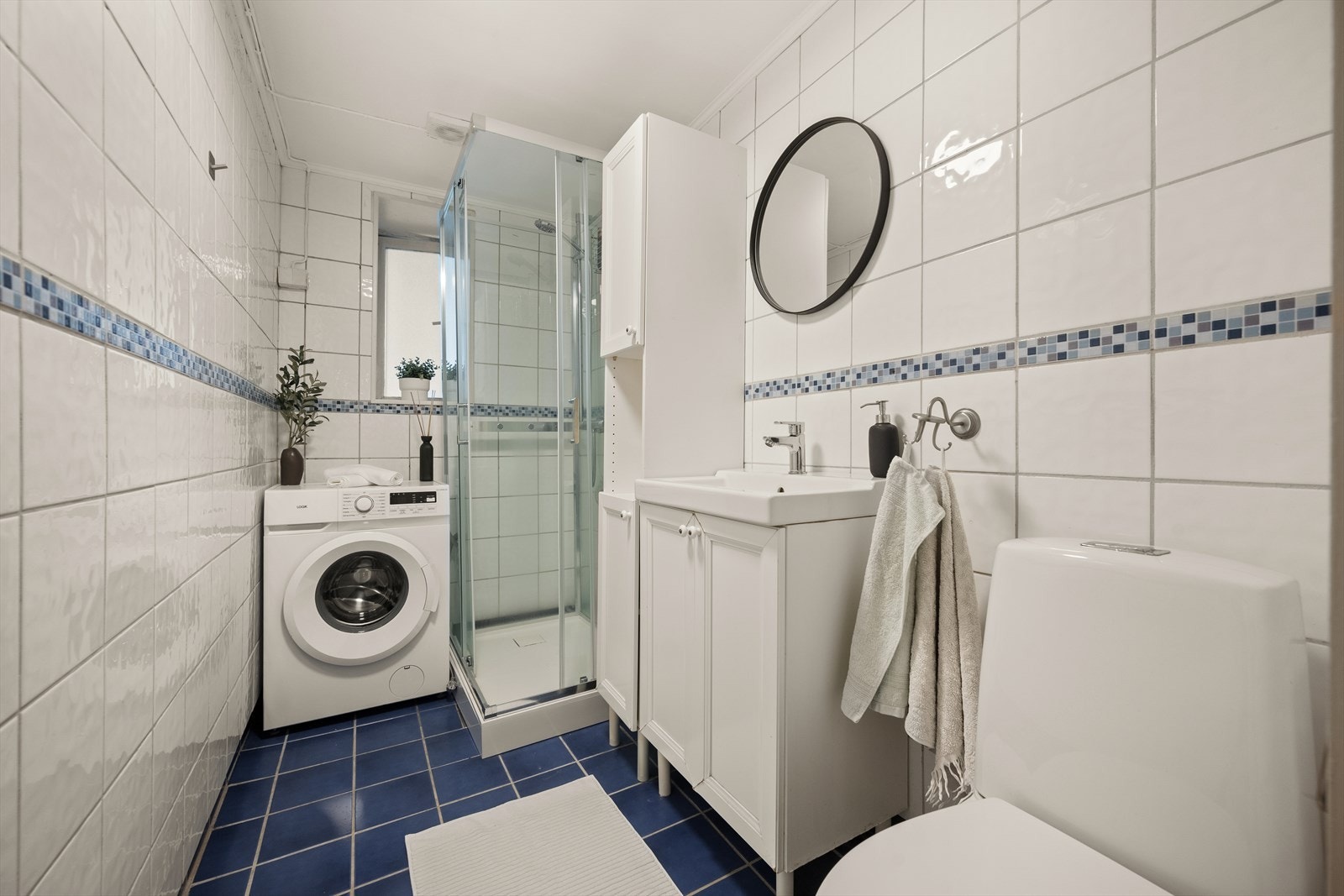 Bad med flislagte overflater, elektrisk styrt vifte og opplegg for vaskemaskin. Badet er innredet med klosett, servant nedfelt i innredningen og dusjkabinett. Galleribilde