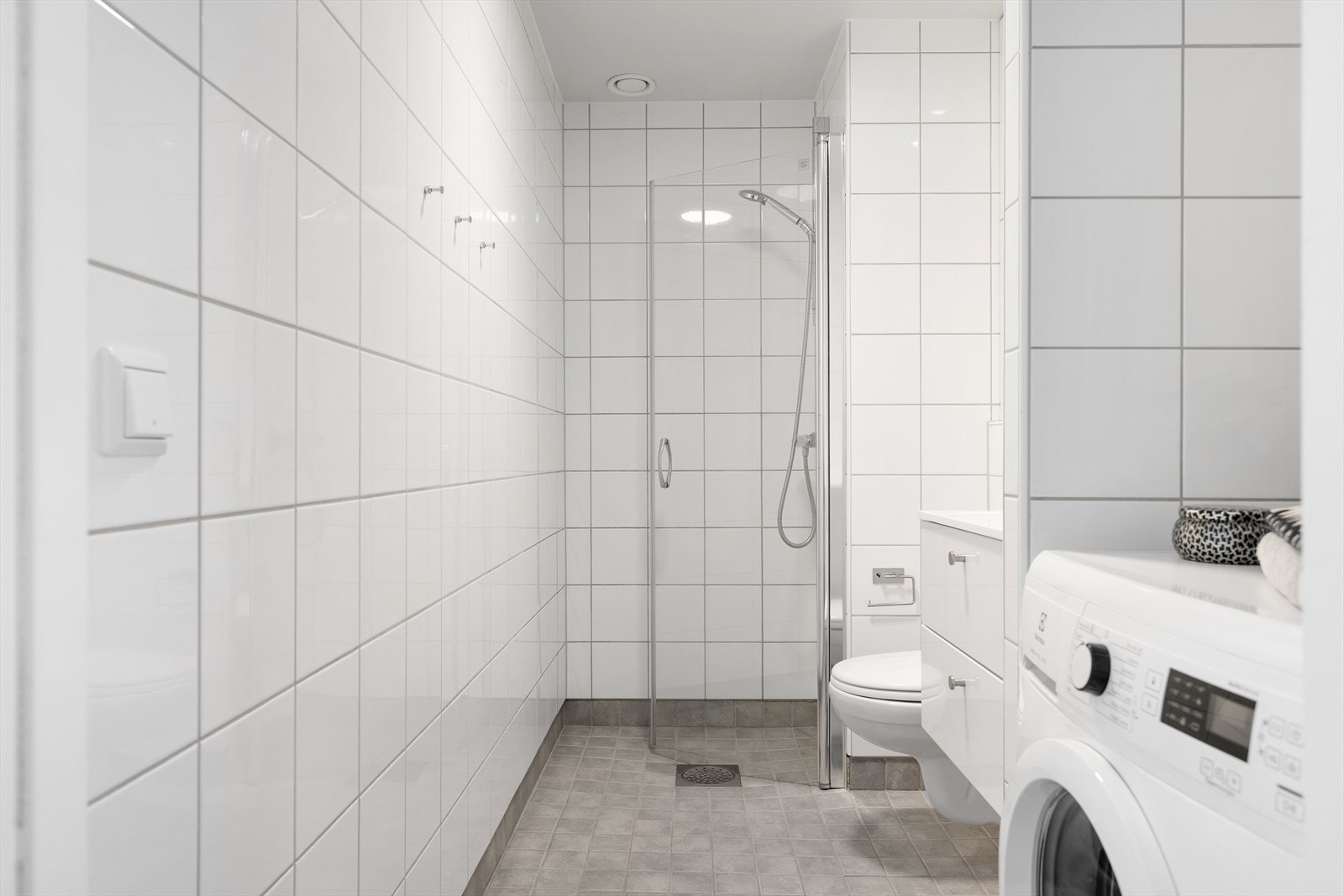 Pent og flislagt baderom med gulvvarme og downlights i himling. Badet er innredet med servantskap med heldekkende servant, speil på vegg, vegghengt toalett med innebygd sisterne samt dusjhjørne med svingdør og termostatstyrt blandebatteri. Galleribilde