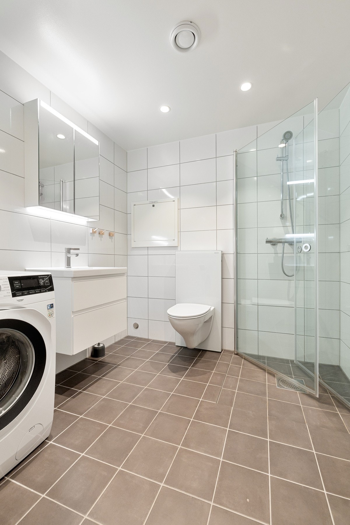 Badet er innredet med nedfelt servant, veggmontert toalett, dusj med glassvegger og opplegg for vaskemaskin. Galleribilde