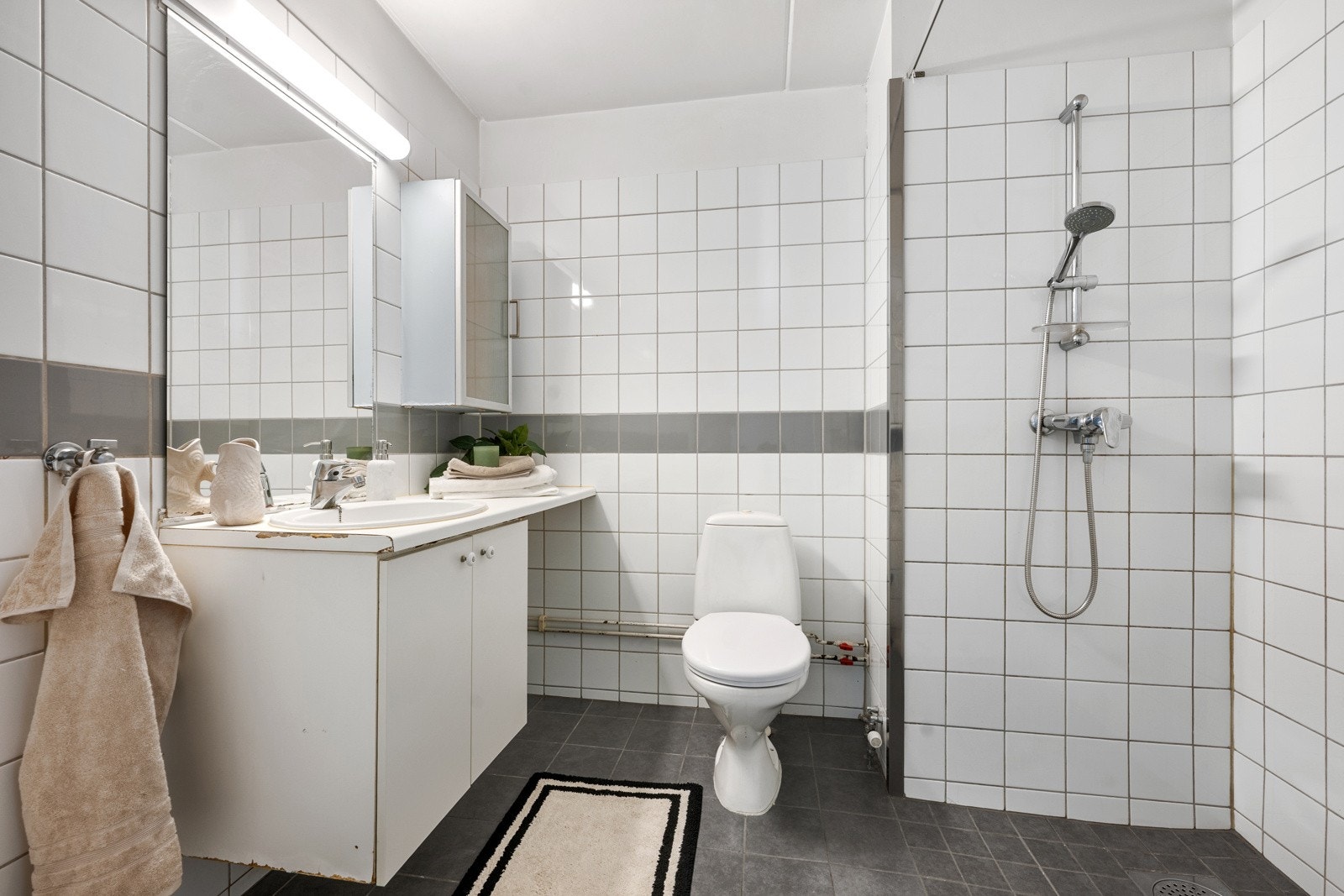 Badet er flislagt og har vannbåren gulvvarme som gir en behagelig temperatur. Rommet er innredet med dusjnisje, servant med underskap, speil med belysning og toalett. Det er i tillegg opplegg for vaskemaskin. Galleribilde