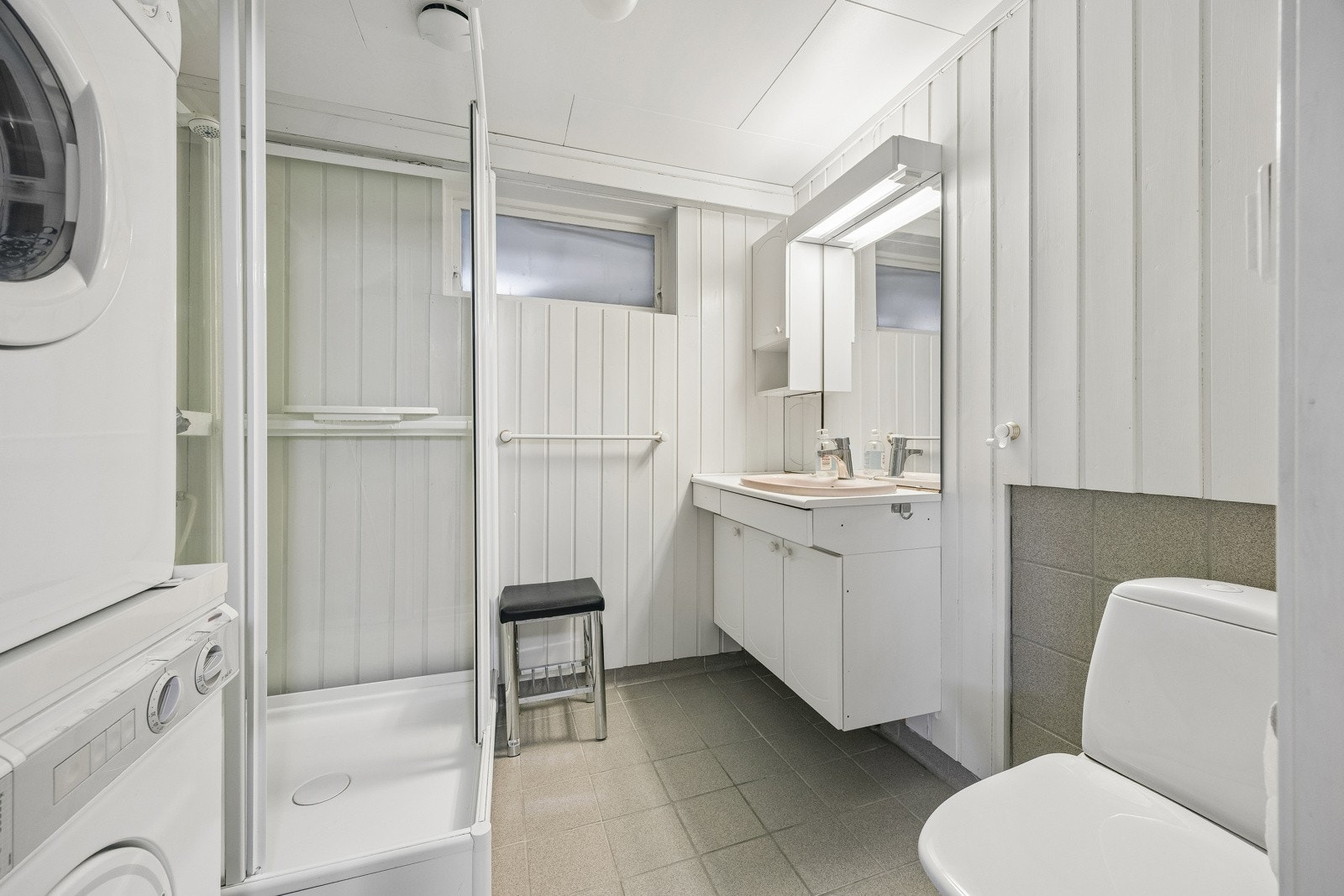 Bad/wc/vaskerom i sokkelleilighet med fliser på gulv og gulvvarme, samt dusjkabinett.