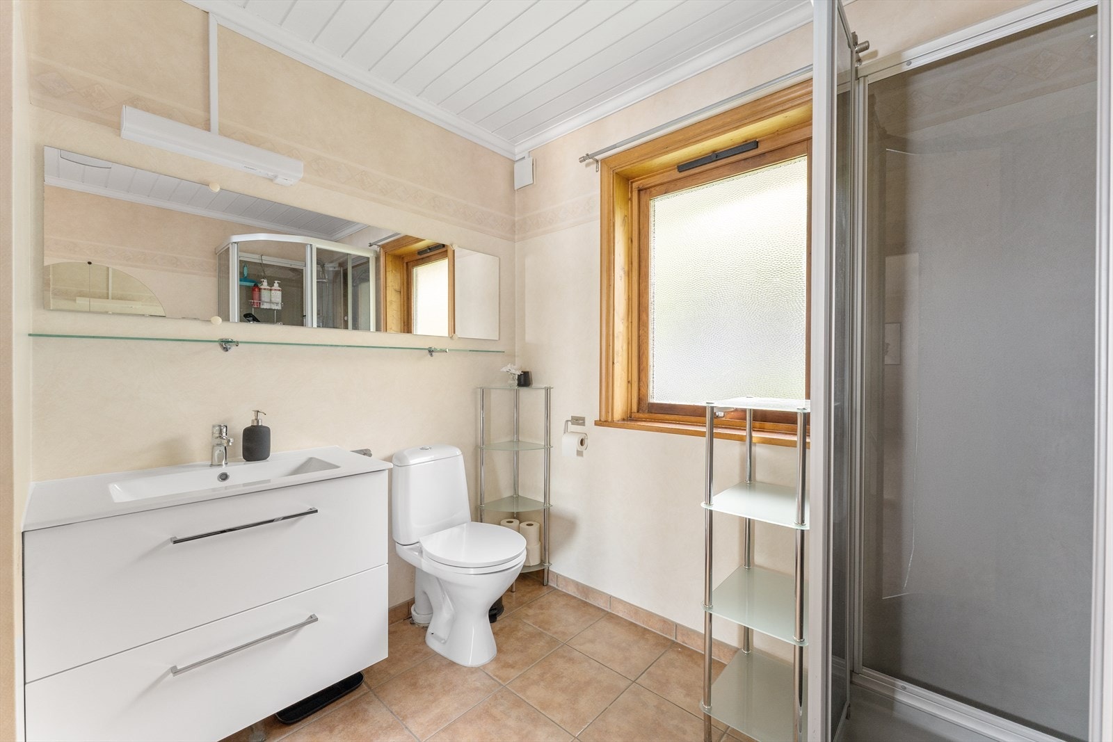 Badet har tapetserte vegger og malt panel i himlingen, samt servant med underskap og høyglansfronter, speil med belysning, dusjkabinett og gulvmontert toalett. Galleribilde