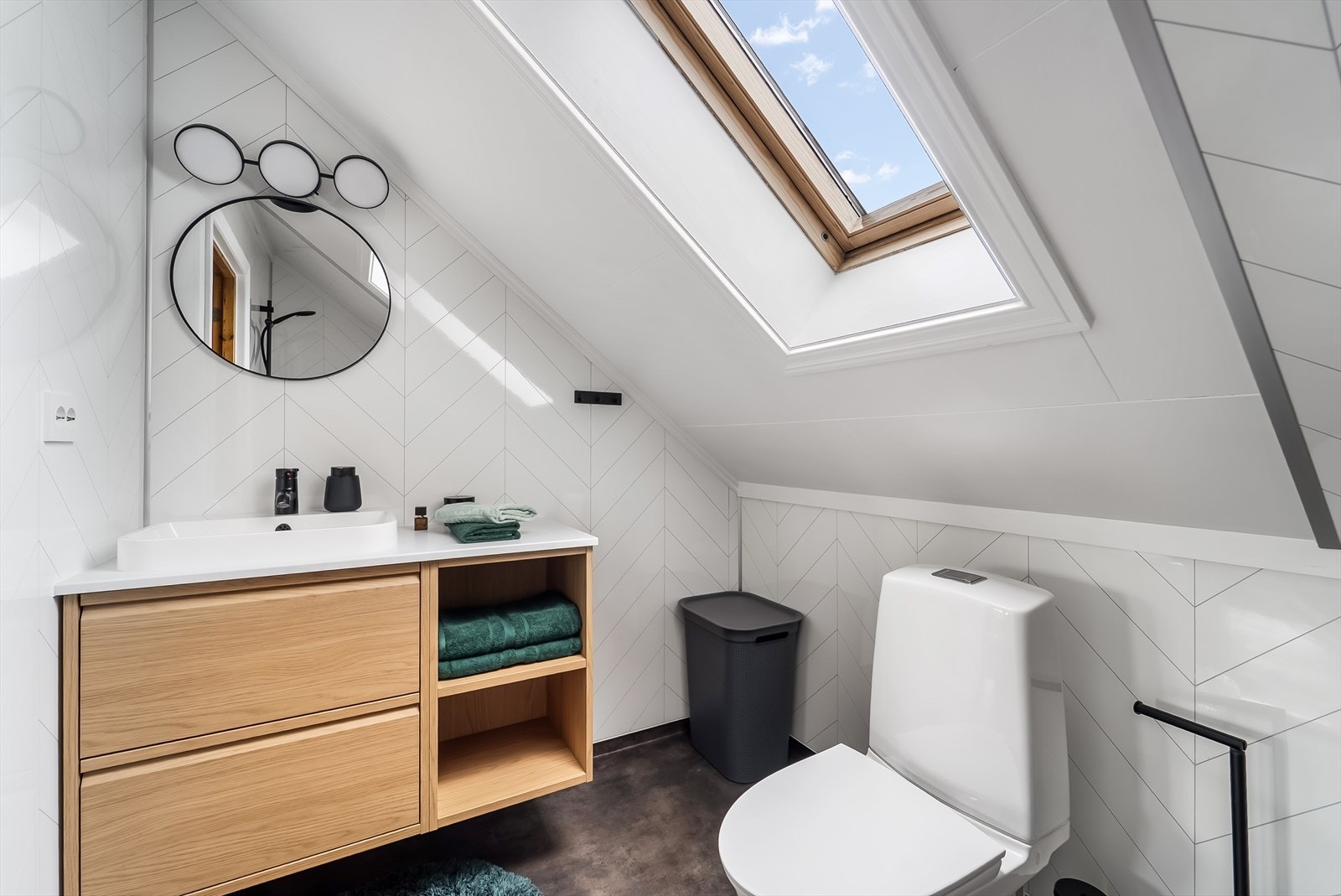 Badet i loftsetasjen fremstår moderne og innbydende med lyse flater, tidsriktige materialvalg og takvindu som gir rikelig med naturlig lys. Rommet er innredet med dusjsone, toalett og servantseksjon med oppbevaring. Galleribilde