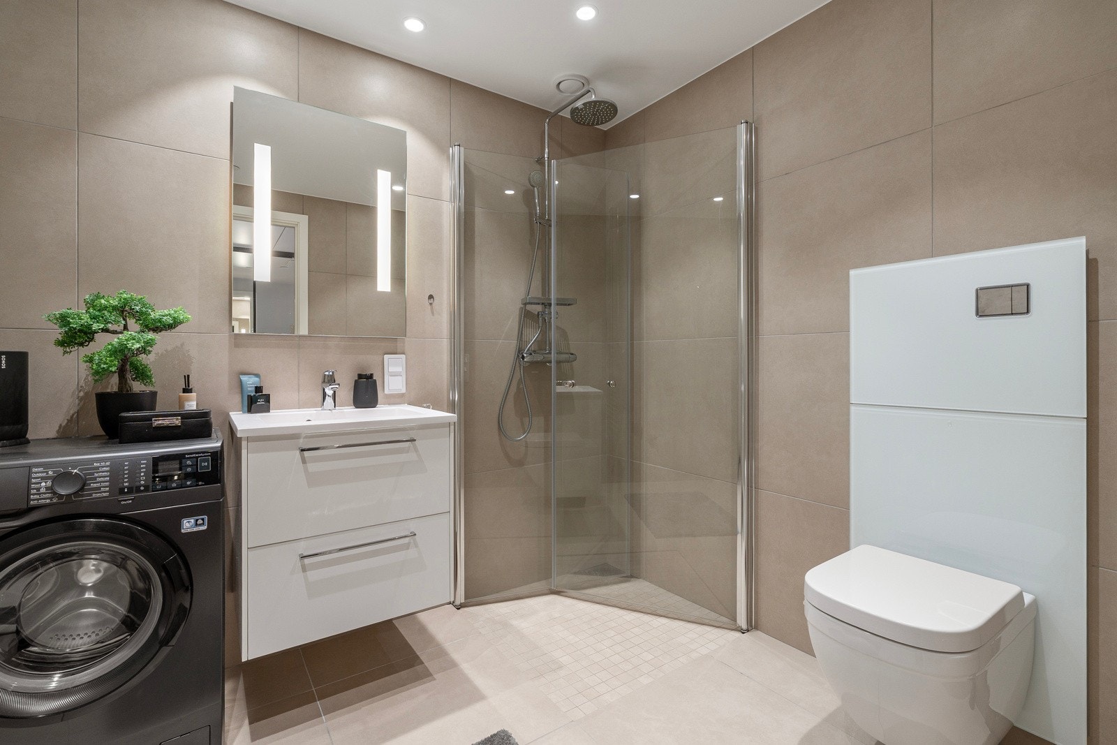 Badet framstår lekkert med store fliser og moderne innredning. Stort dusjhjørne med innfellbare glassdører, praktisk vegghengt wc og plass til vaskemaskin