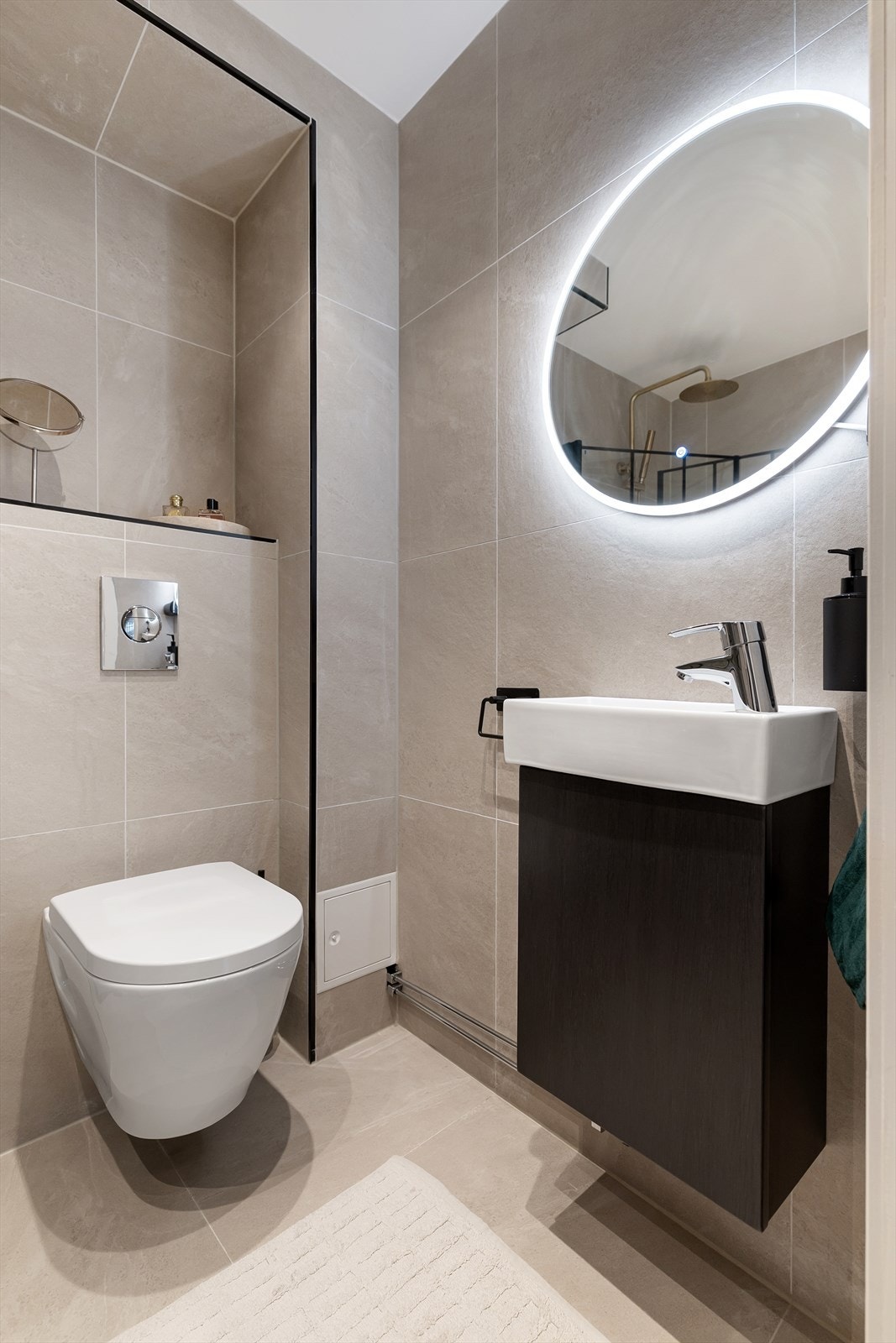 Badet er utstyrt med dusjhjørne med innfellbare dusjdører, veggmontert WC og servant med praktisk underskap. Over servanten er det et rundt speil med integrert belysning, som gir et moderne og elegant uttrykk. Galleribilde