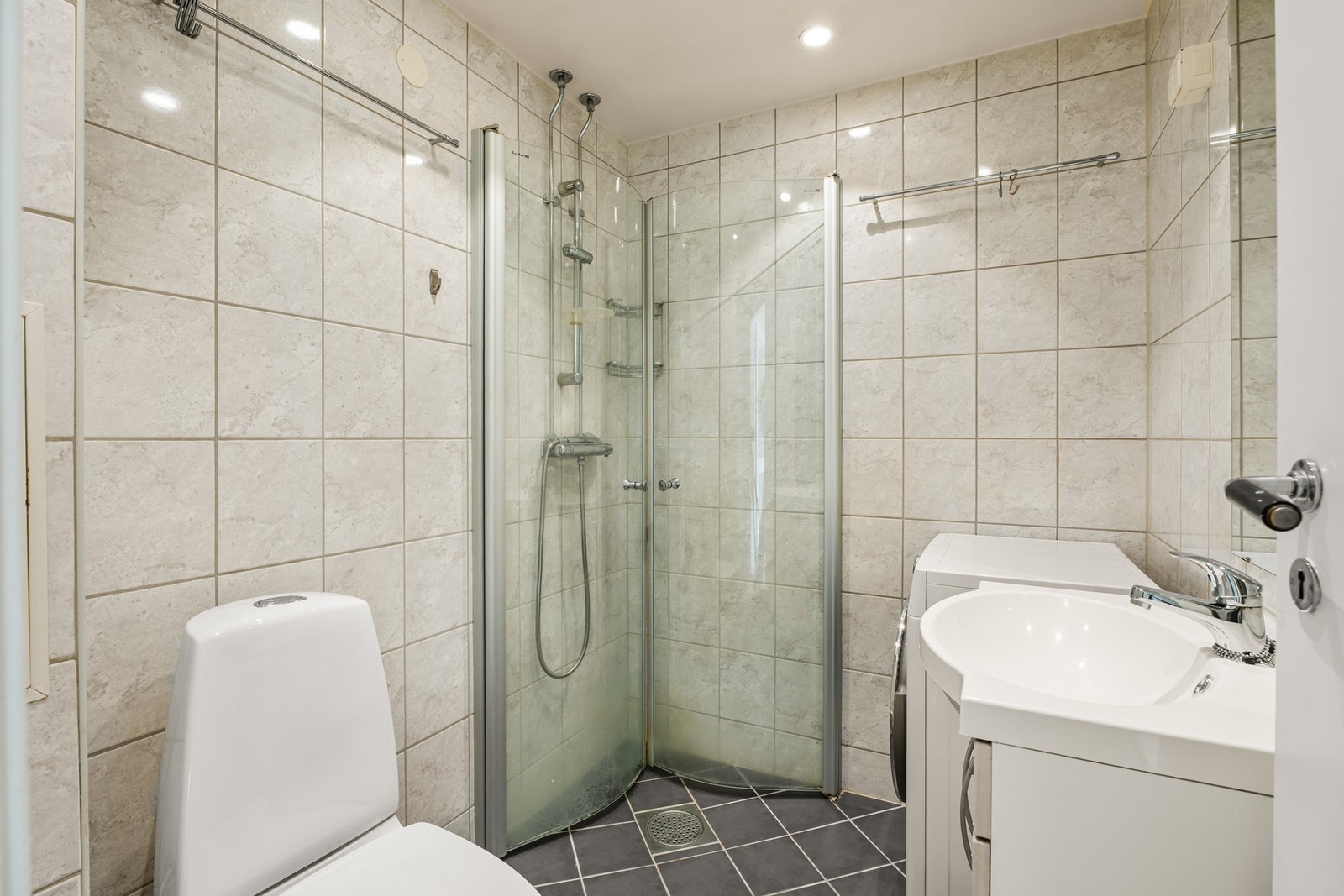 Badet har vegghengt servant og underliggende innredning med to skapdører av slette fronter. Dusjsonen er adskilt med to skyvbare buede glassdører. Det er opplegg for vaskemaskin. Galleribilde