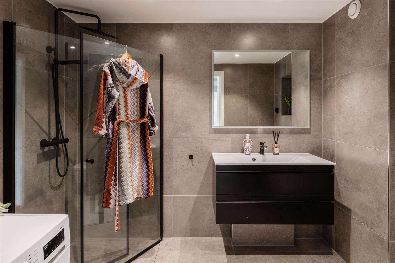 Badet ble smakfullt pusset opp i 2021 og har bl.a. opplegg for vaskemaskin, fliser på gulv og vegger, samt gulvvarme og downlights. Alle detaljer er holdt i et trendy, sort design. I 2024 ble det også montert et dobbelt støpsel. Galleribilde
