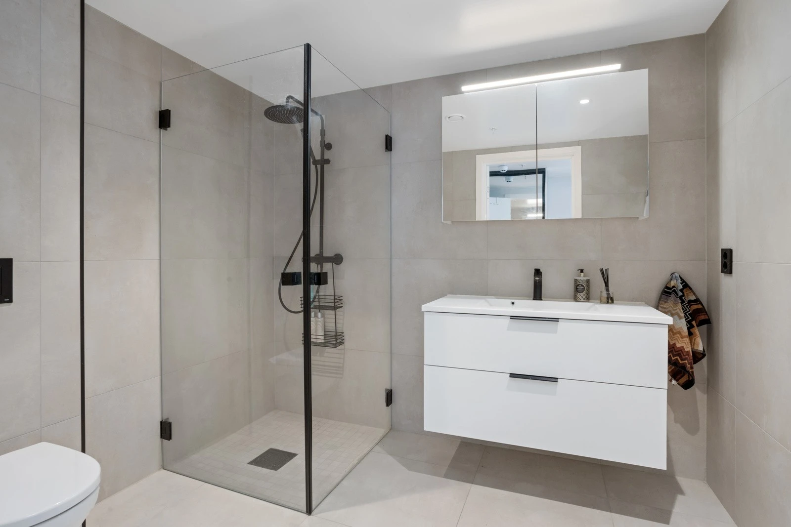 Delikat og påkostet baderom med storformatet 60x60 fliser. Pen baderomsinnredning med sorte detaljer, veggheng toalett, servant med dobbel skuff, speilskap med belysning over, sort armatur og dusjhjørne med glassdører Galleribilde