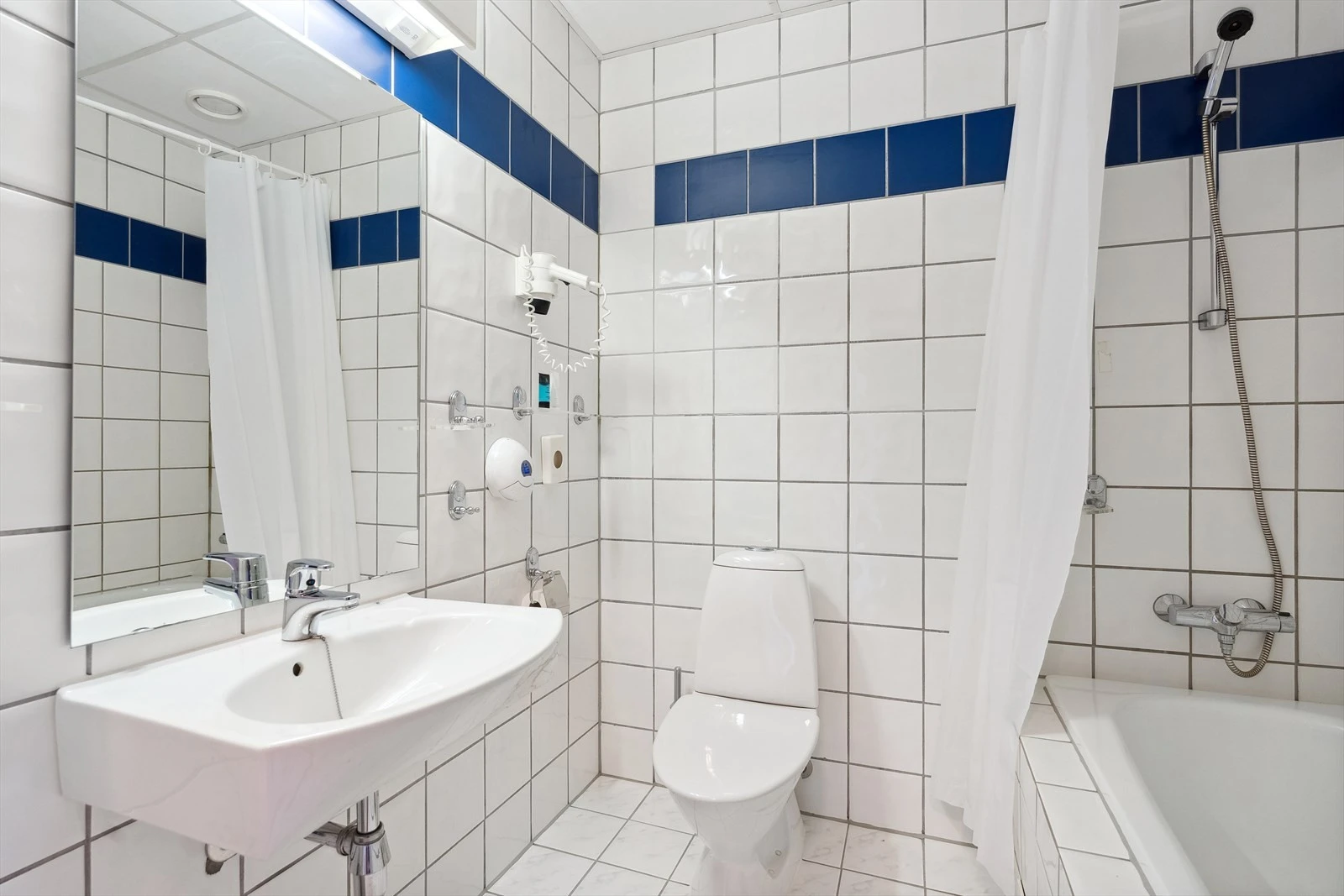 Bad med flisbelagte overflater, med varmekabler i gulvet. Det er montert vegghengt porselenservant, gulvstående toalett, vegghengt speil og badekar med dusjgarnityr på vegg. Galleribilde